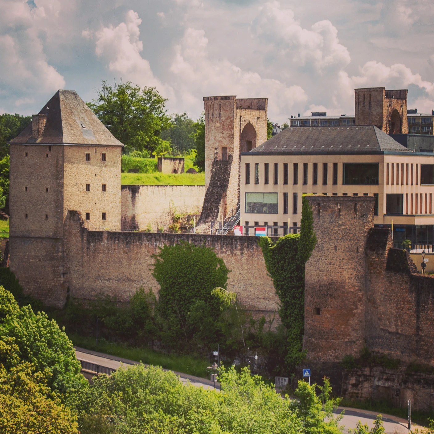 Stone towers and walls, partly covered with greenery with a modern building in the center in Luxembourg City.