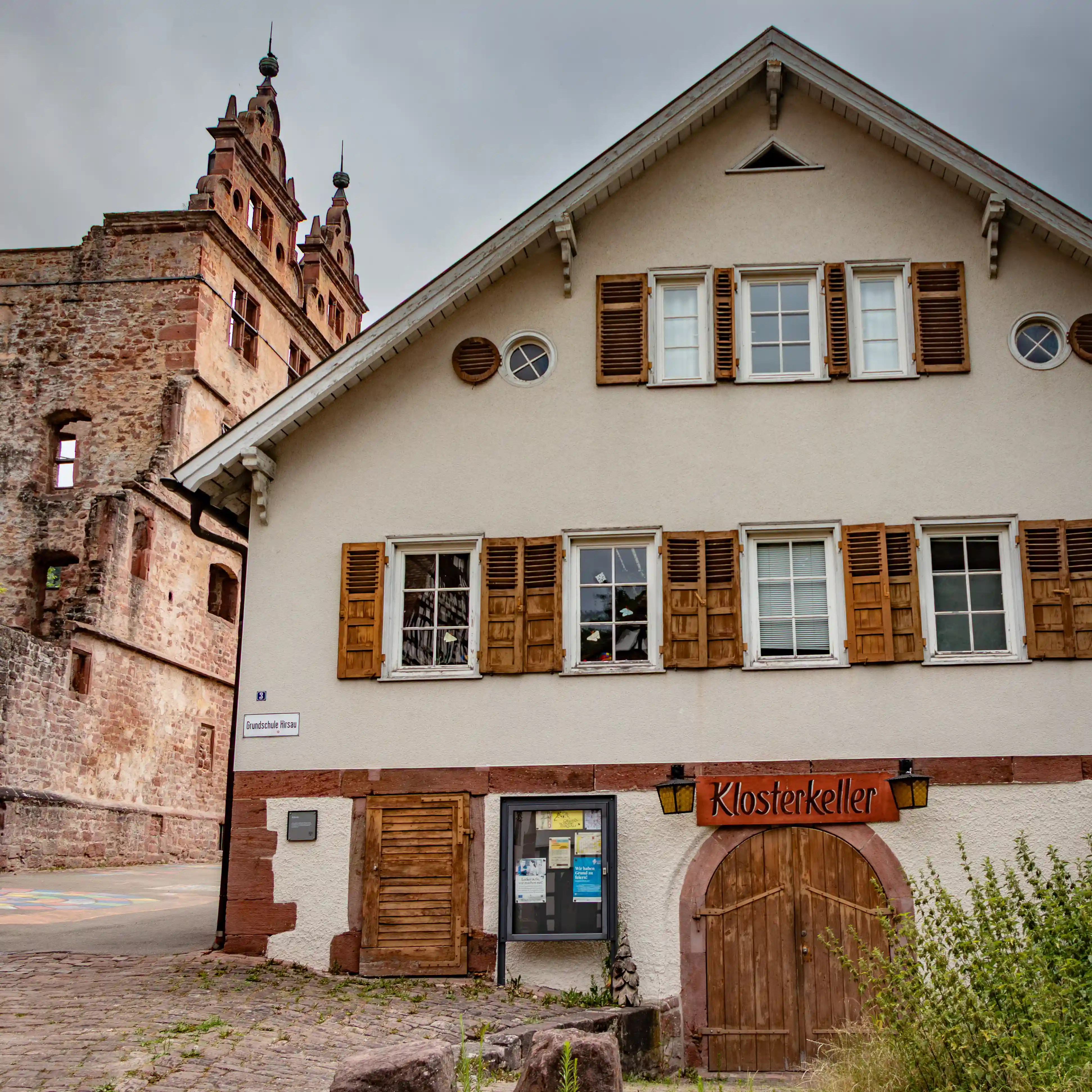 White plastered building with wooden shutters marked “Klosterkeller,” beside the Hunting Lodge ruins in Hirsau.
