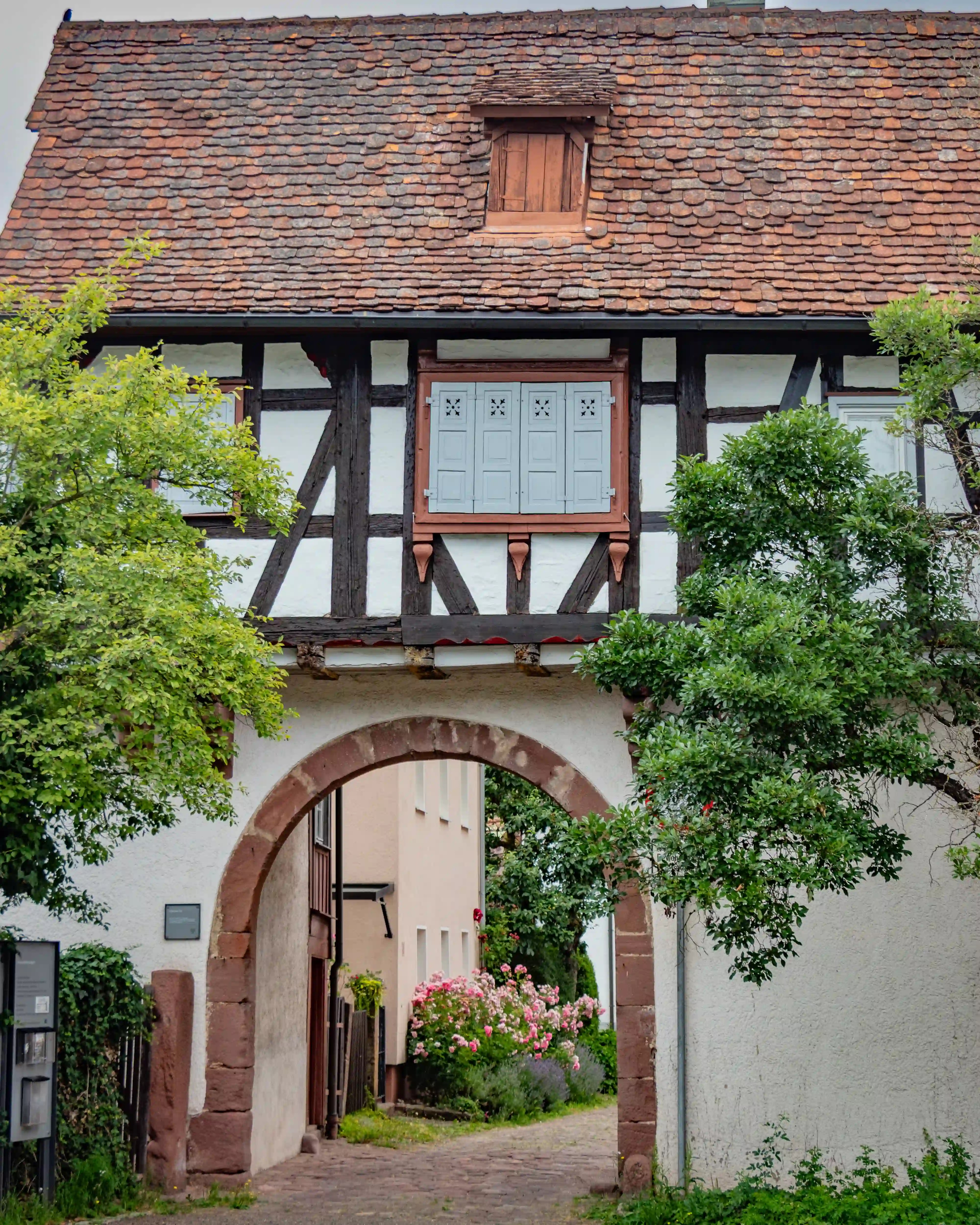 Half-timbered building with an archway at Hirsau Monastery, framed by trees and blooming flowers.