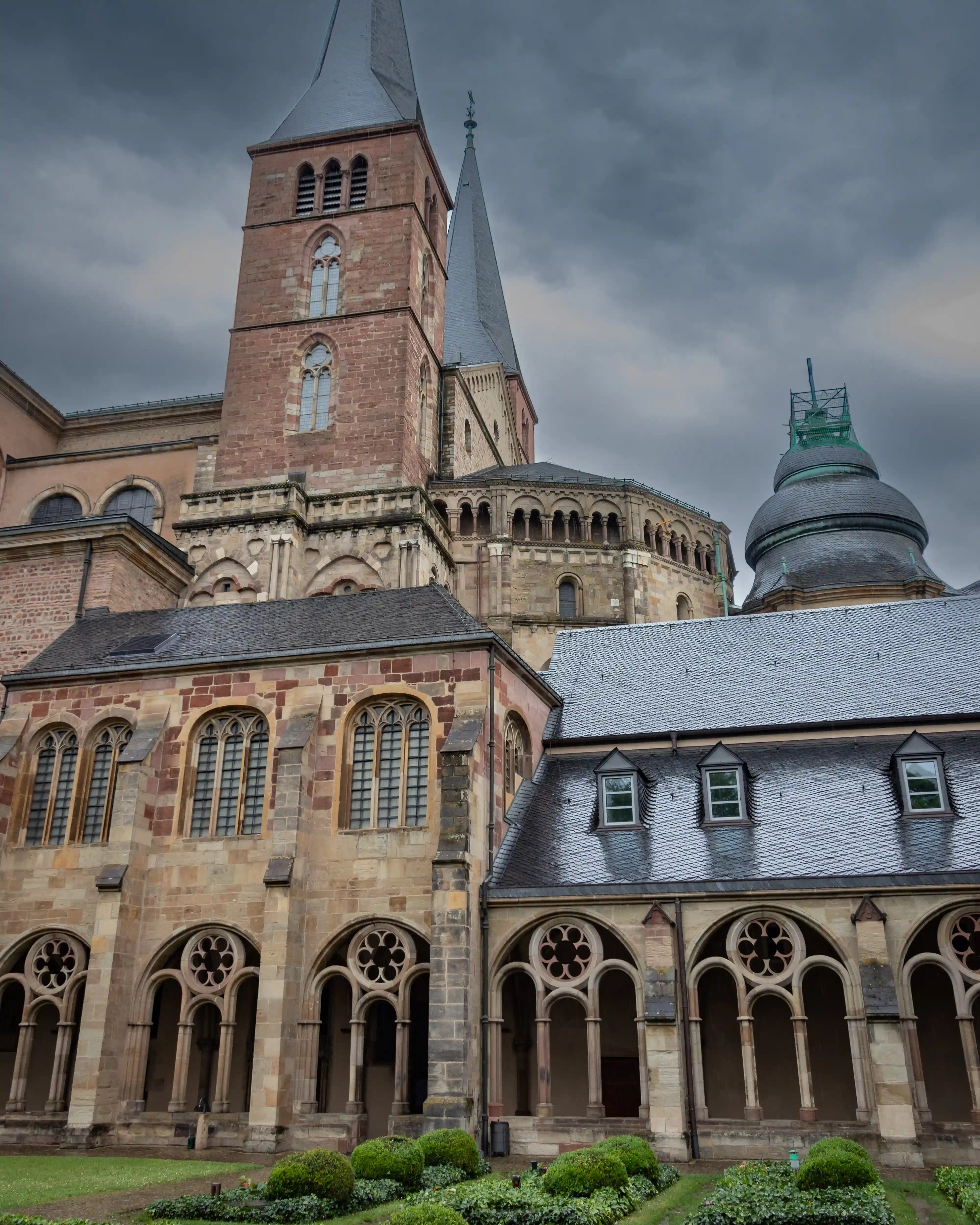 Exterior of Trier Cathedral with Romanesque towers and the adjacent cloister garden under cloudy skies.