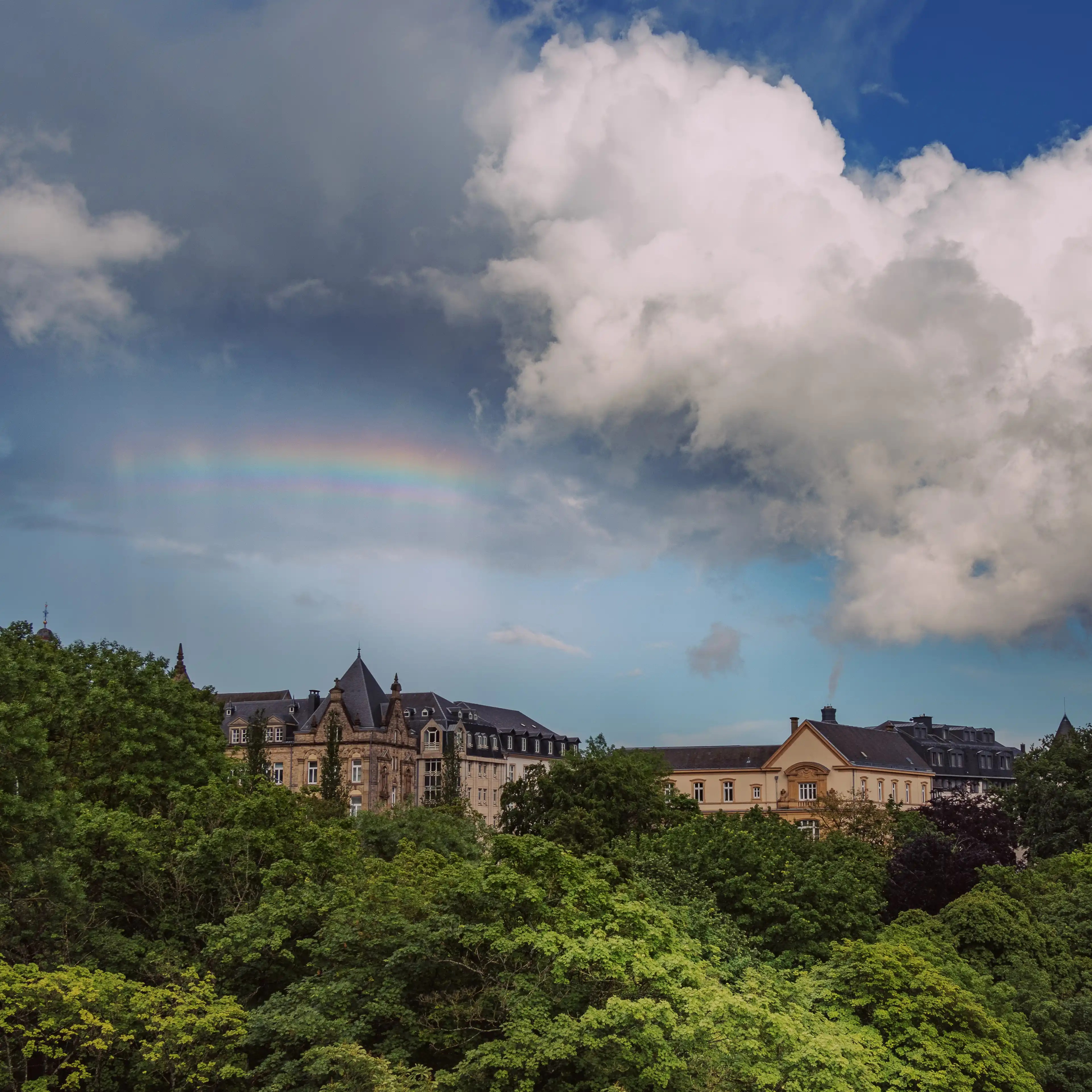 A faint rainbow arcing across the sky above historic buildings and green treetops in Luxembourg City.