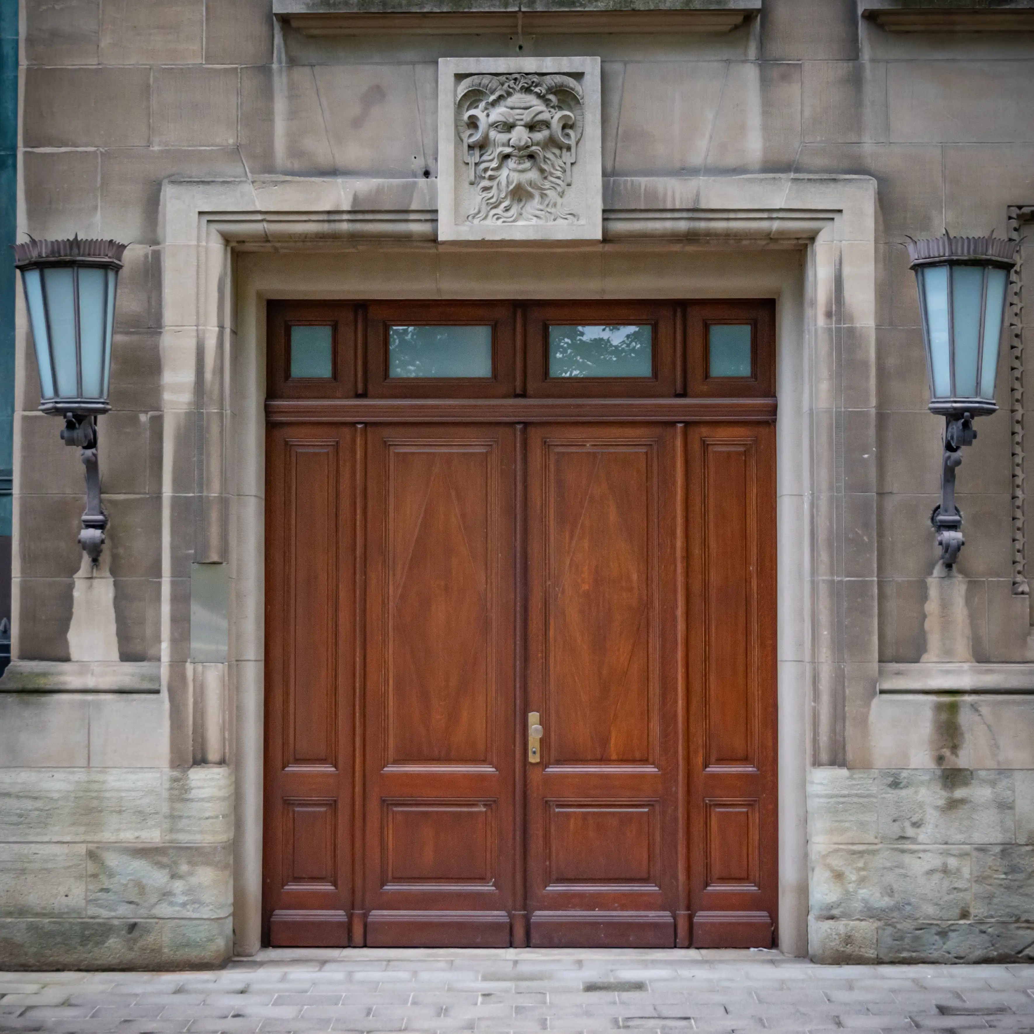 Large wooden double doors framed in stone, with two lantern-style lights on either side and a carved face above the entrance.