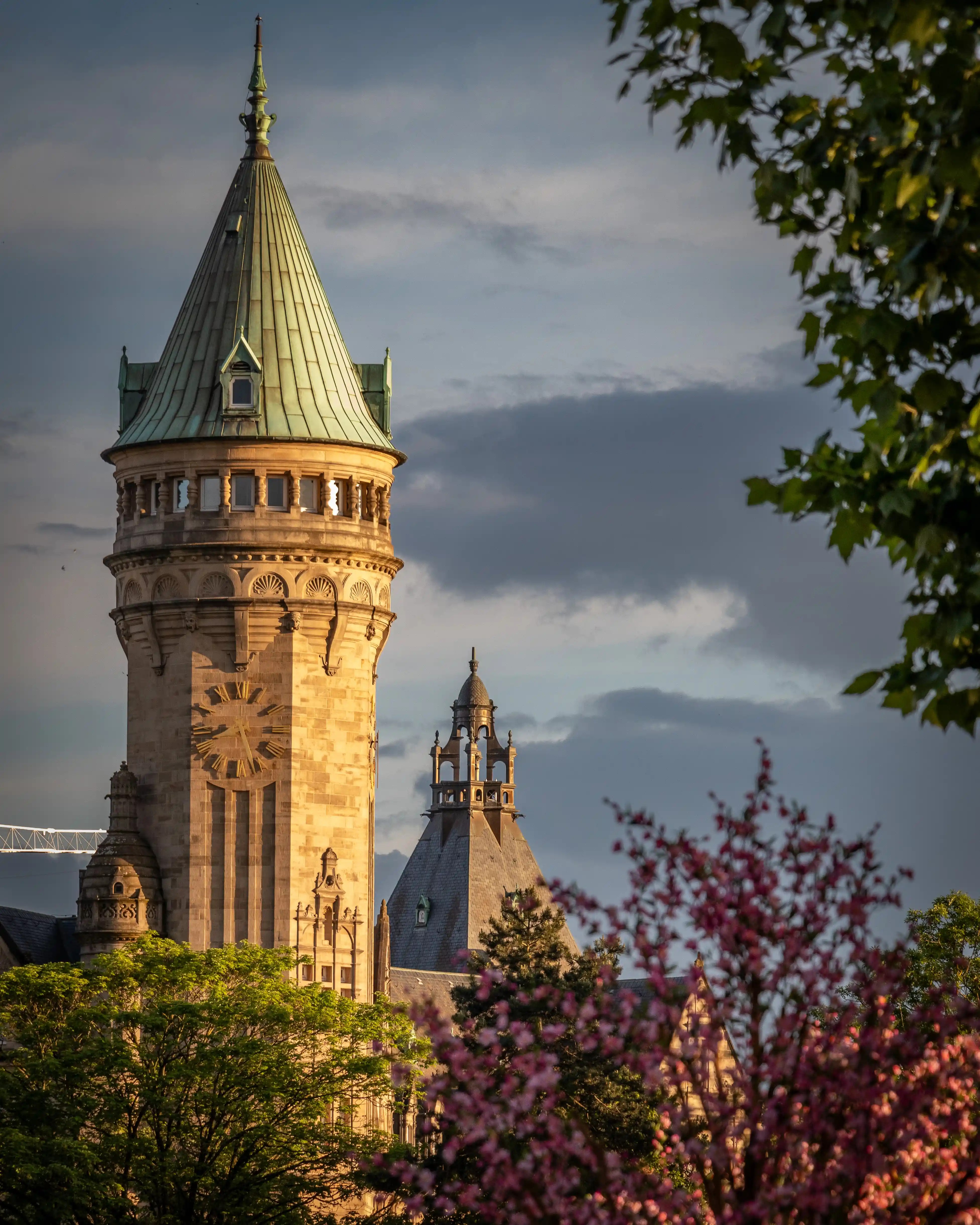 Tower of the Luxembourg City bank building with a green copper roof, clock face, and detailed stonework, framed by trees and evening light.