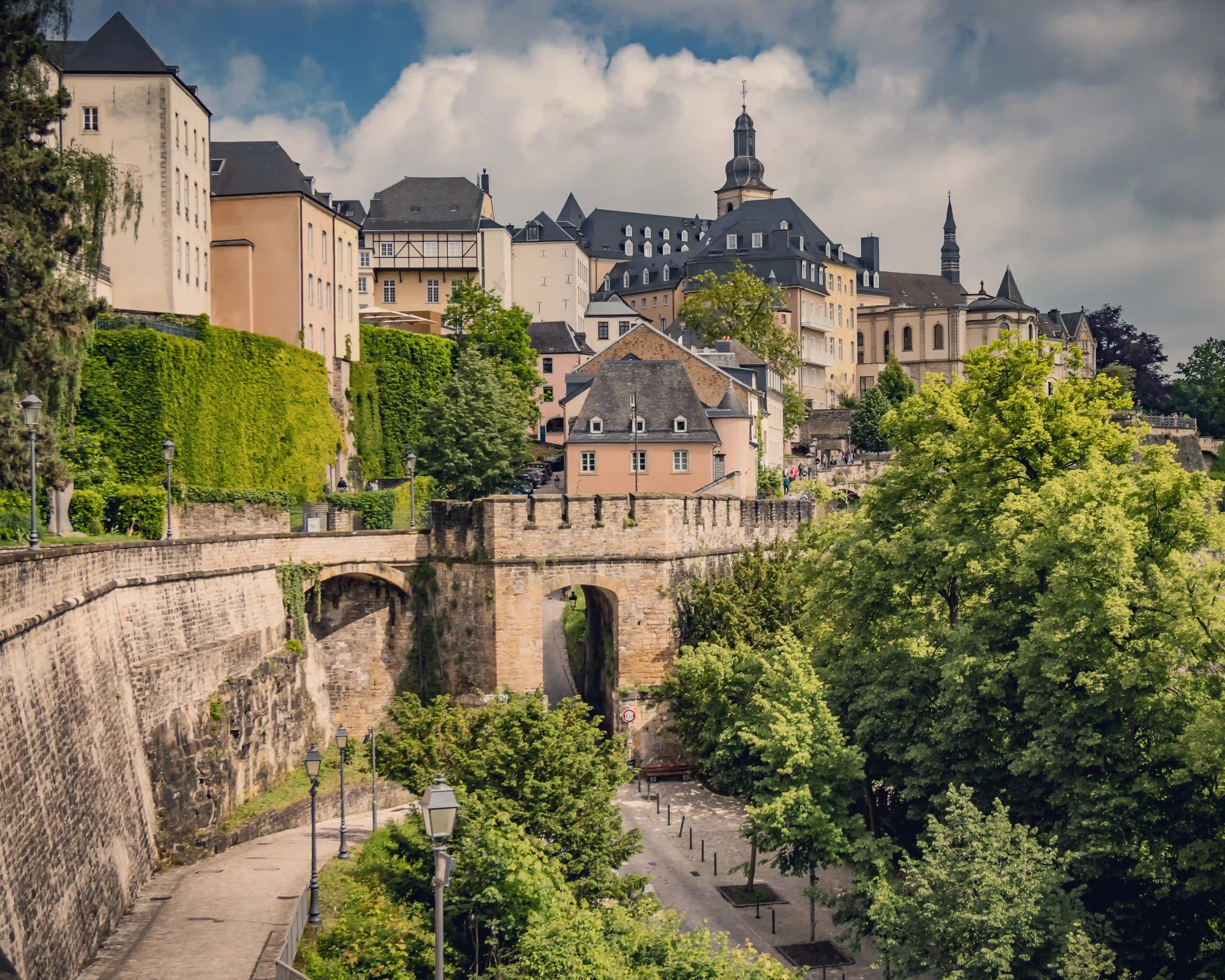 View of Luxembourg City’s historic fortifications with stone walls, arched bridge, and pastel buildings rising above, framed by greenery.