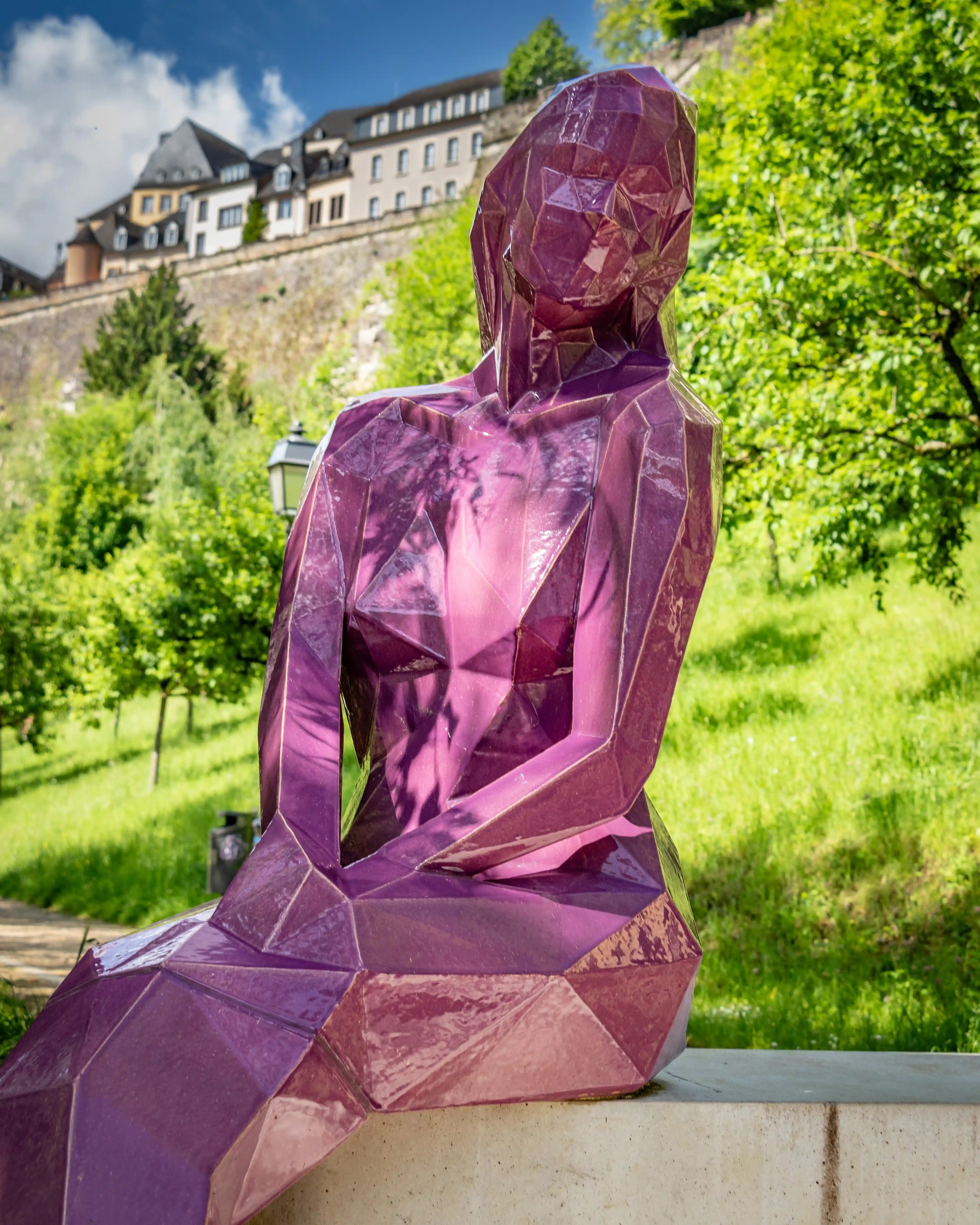A modern geometric purple statue of Melusina, the legendary mermaid of Luxembourg, seated with her tail folded, set against greenery and the old city walls in the background.