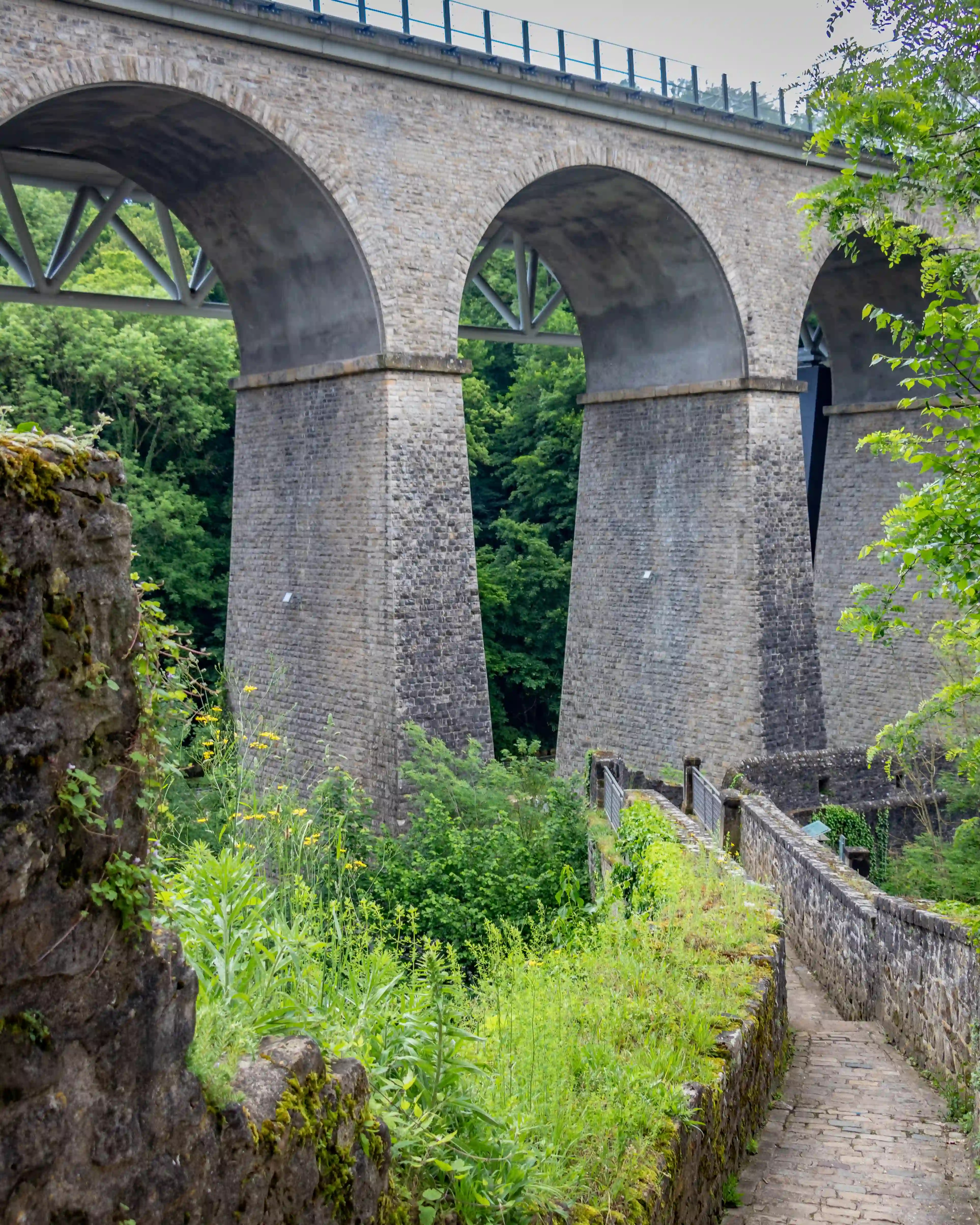 Stone railway viaduct with tall arches surrounded by greenery, with a small cobblestone path in the foreground.