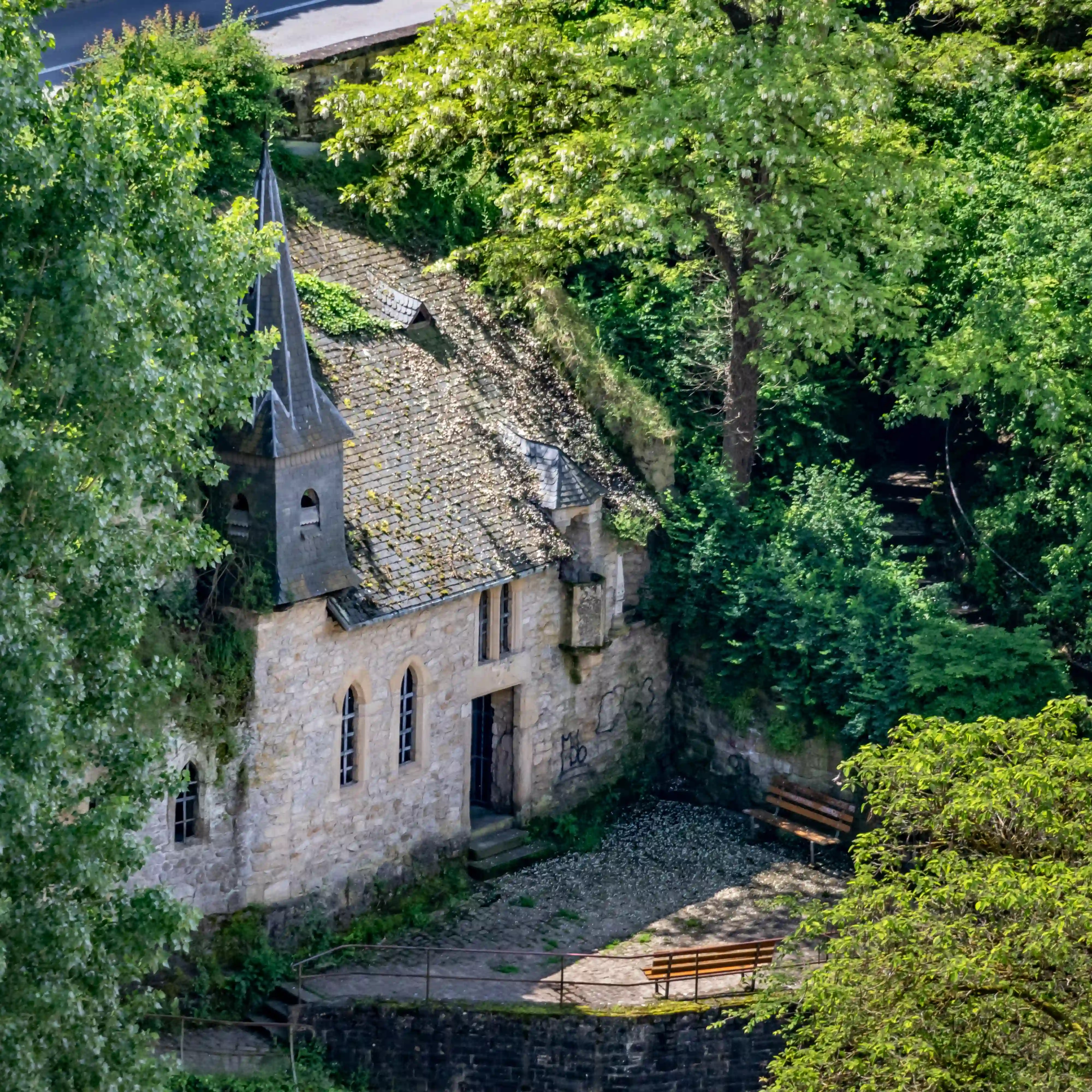 Small stone chapel with a steep roof and pointed bell tower, partly hidden by tall trees in Luxembourg City.