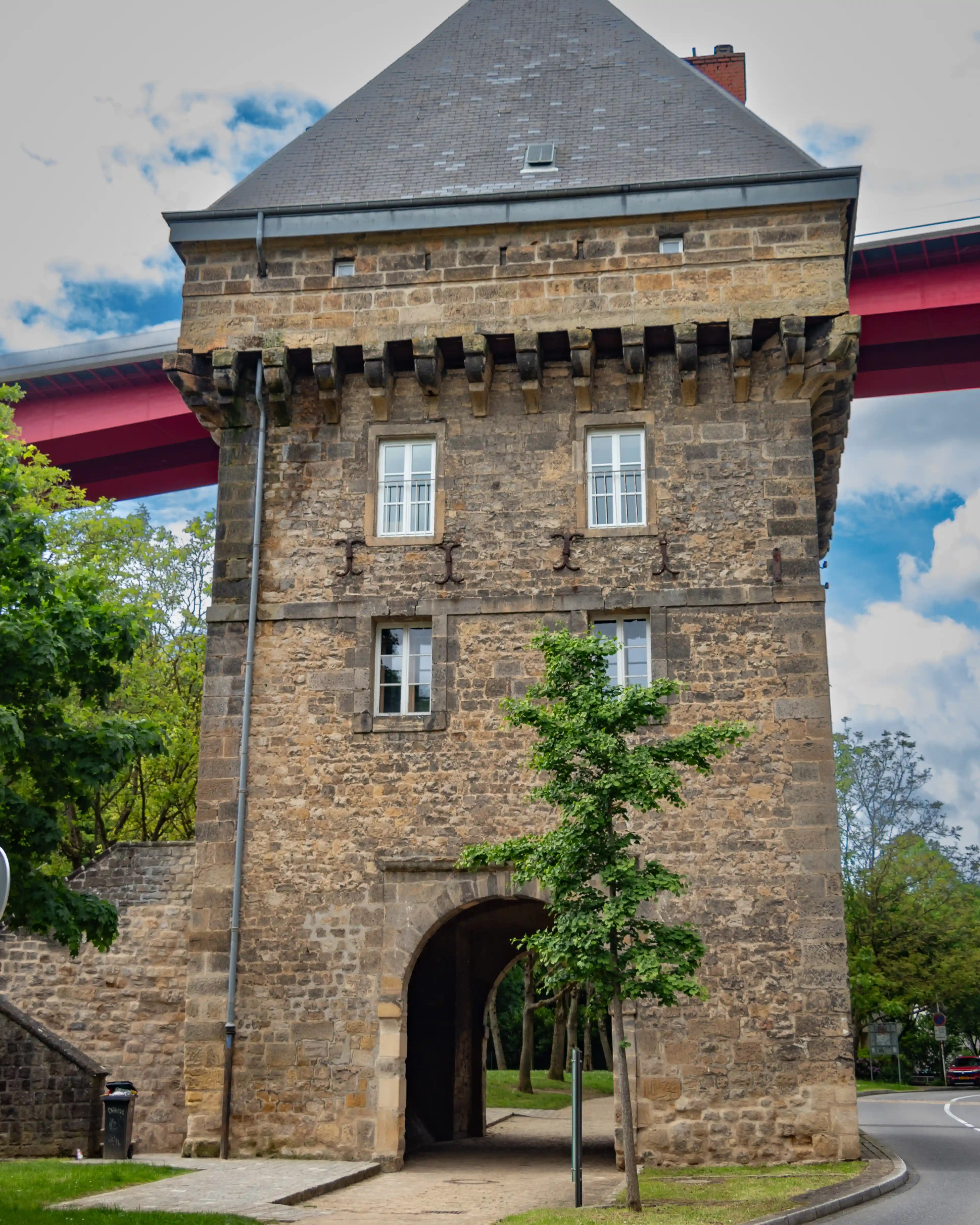 Medieval stone tower with arched gateway beneath the modern red Grand Duchess Charlotte Bridge.