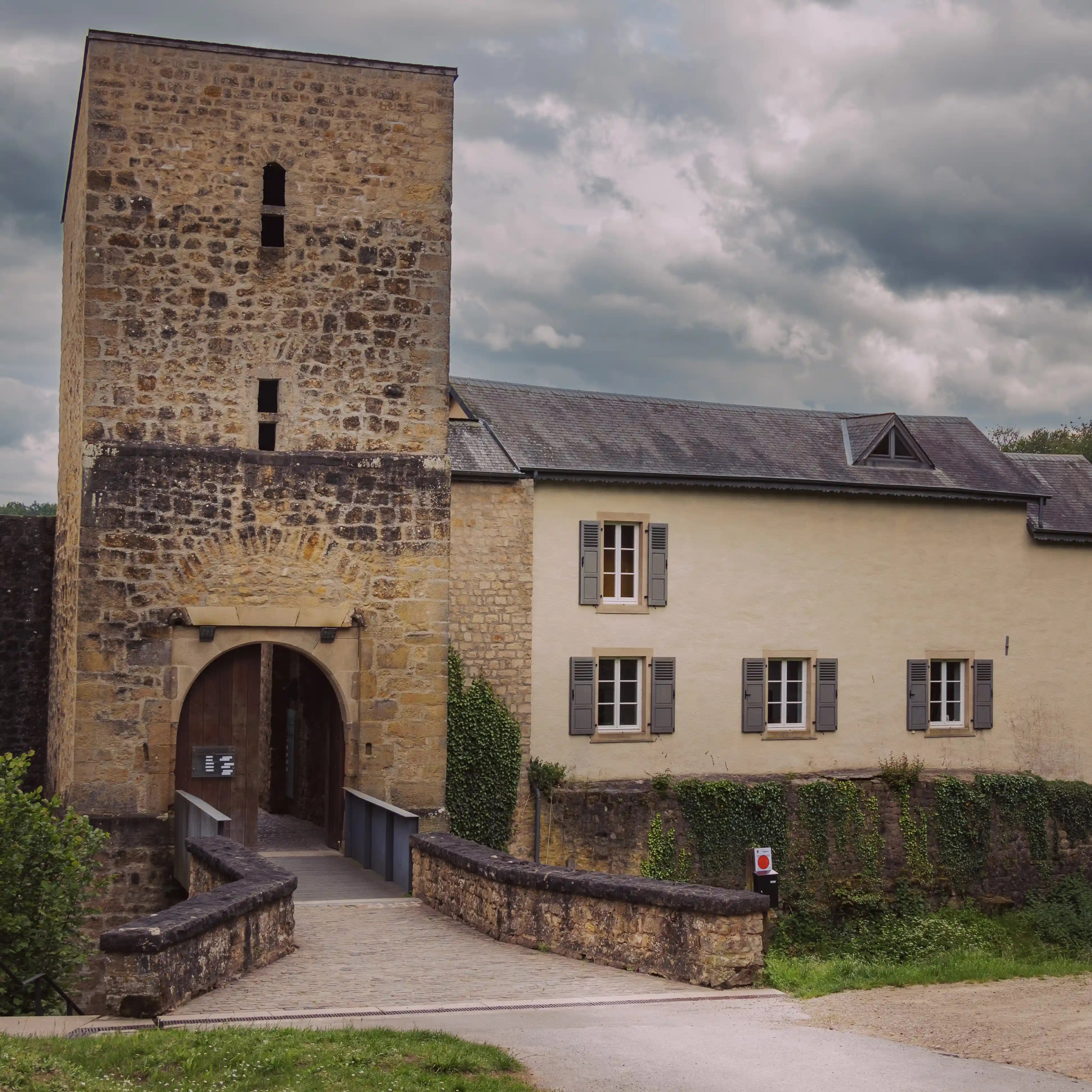 Stone gatehouse of Larochette Castle with arched entrance, flanked by a cream-colored building and a cobblestone path leading across a bridge.