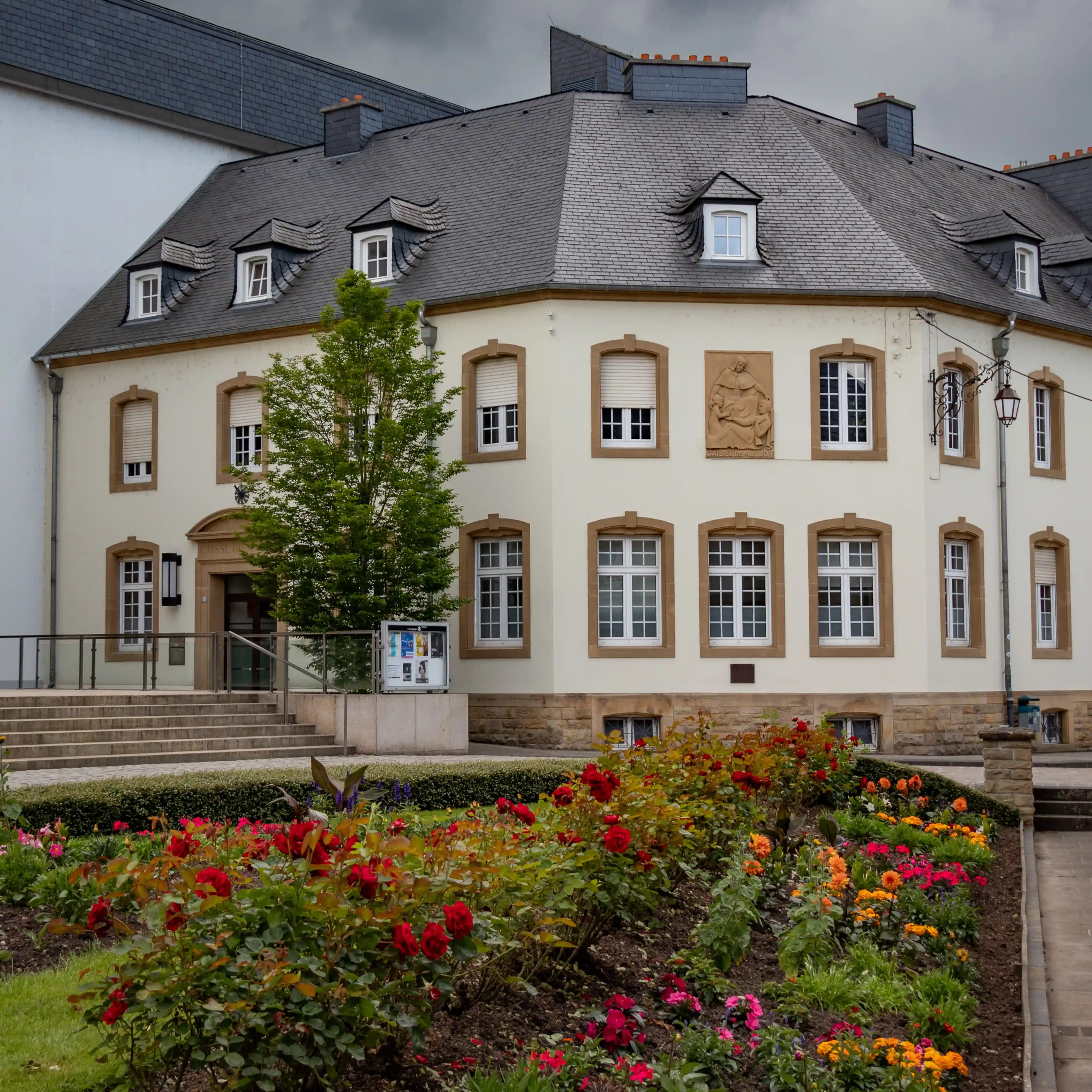 Historic building with cream-colored facade, slate roof, and a front garden full of colorful flowers in Echternach.