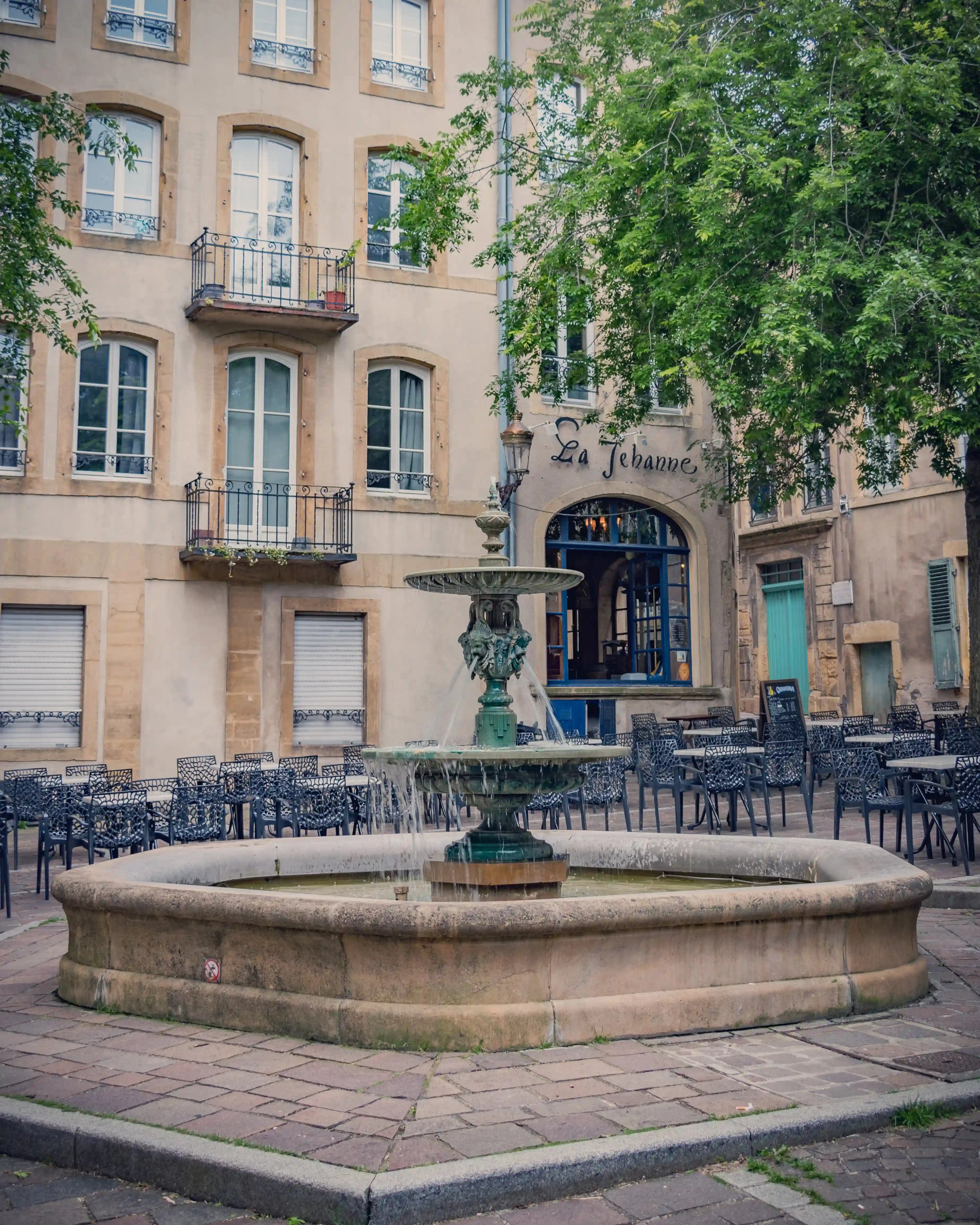 Ornate fountain in a cobblestoned square in Metz, with empty café chairs and the restaurant La Jehanne in the background.