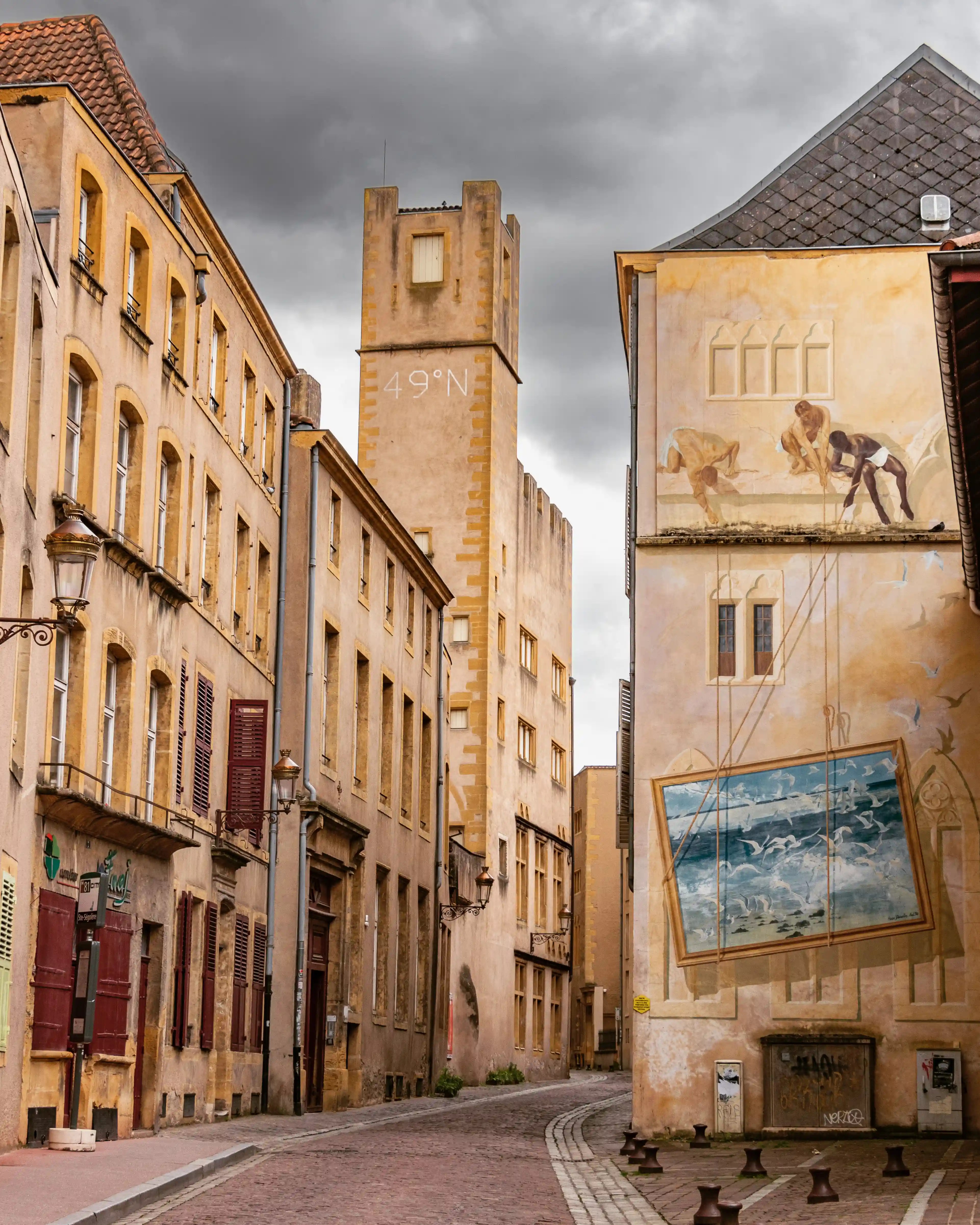 Narrow street in Metz with a painted mural of workers and seagulls, and a tower marked “49°N” rising behind.