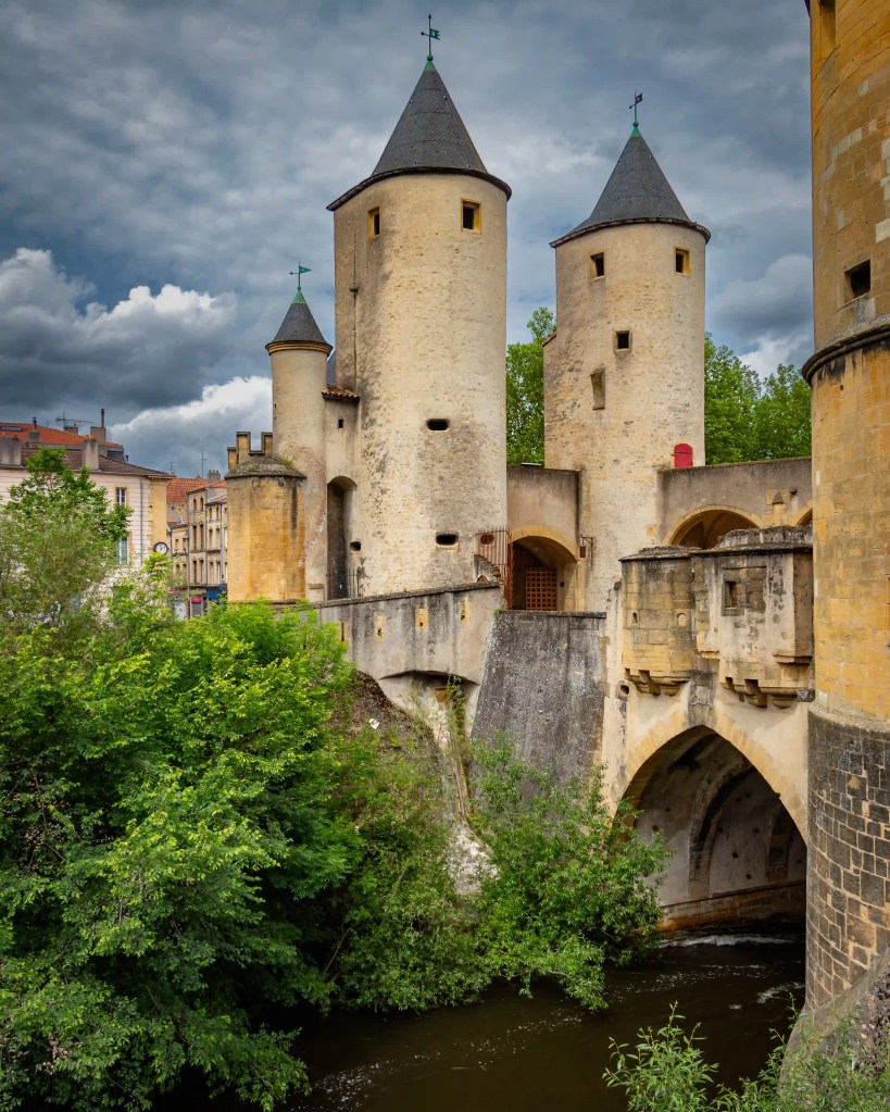 The twin towers of the Porte des Allemands in Metz rising above a stone bridge and river.