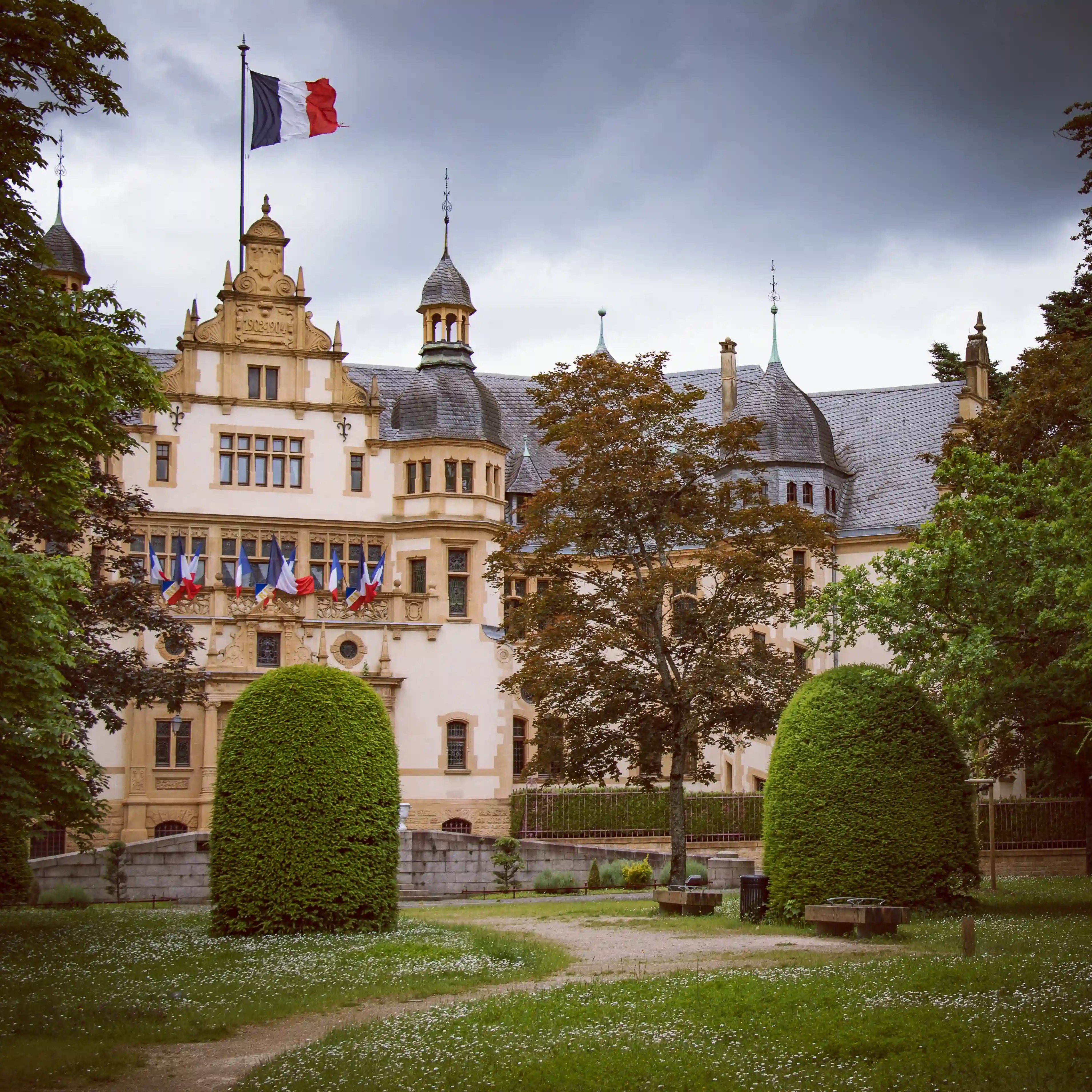 Palais du Gouverneur de Metz with French flags, framed by manicured gardens and trees.