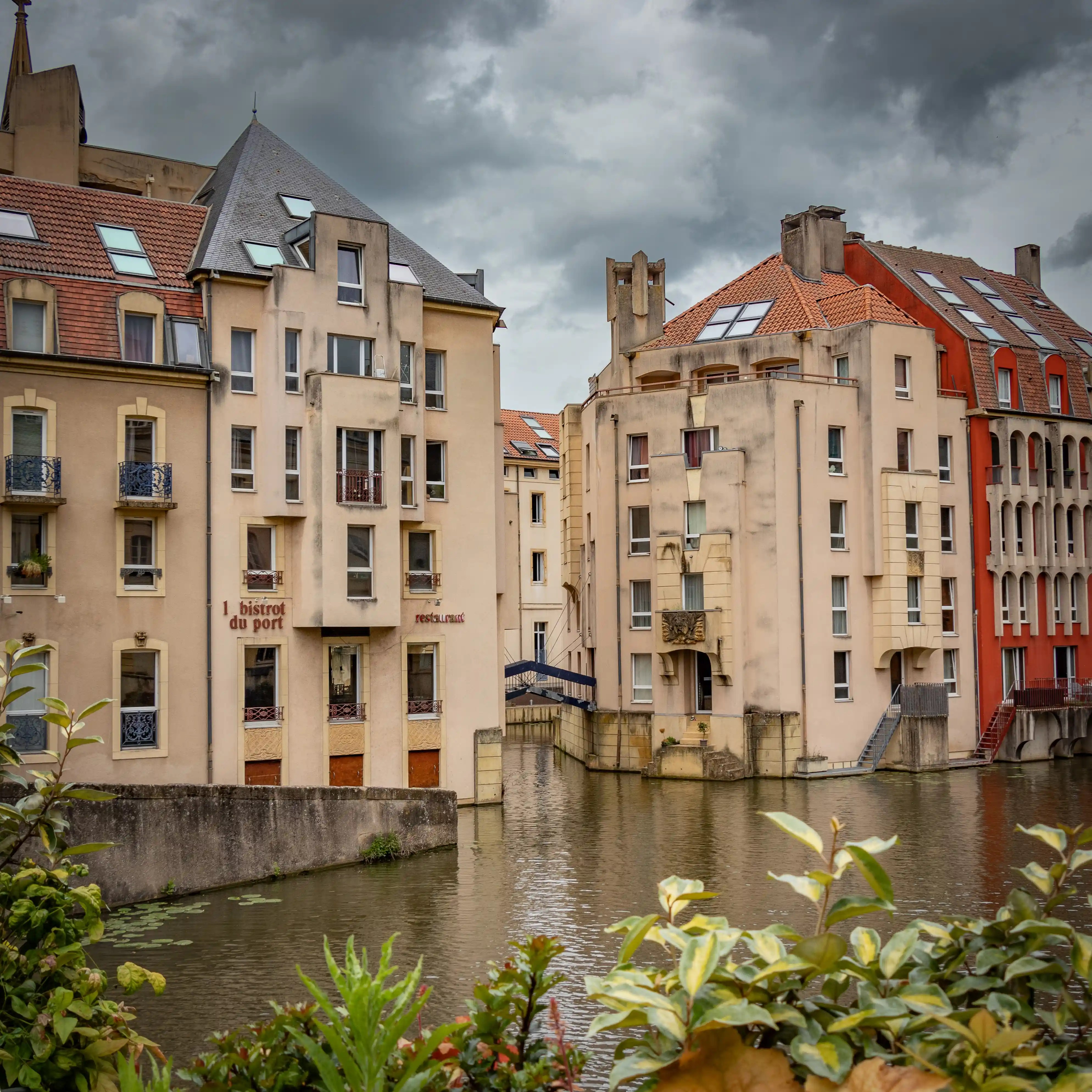 Riverside apartment buildings in Metz, France, with red-tiled roofs and “Le Bistrot du Port” restaurant at ground level, reflected in the Moselle River.