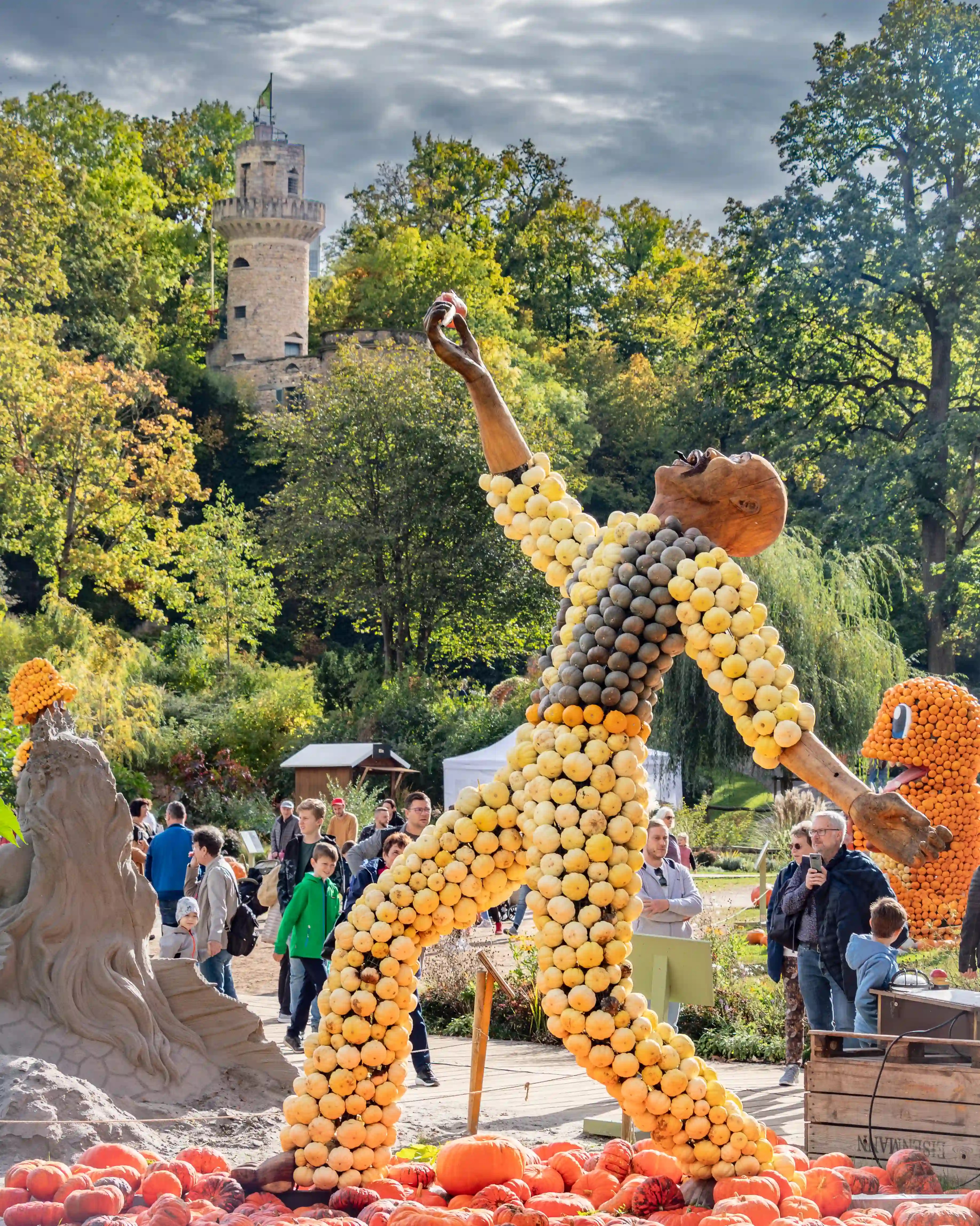 Large pumpkin sculpture of a human figure made from yellow and gray gourds with festival visitors around.