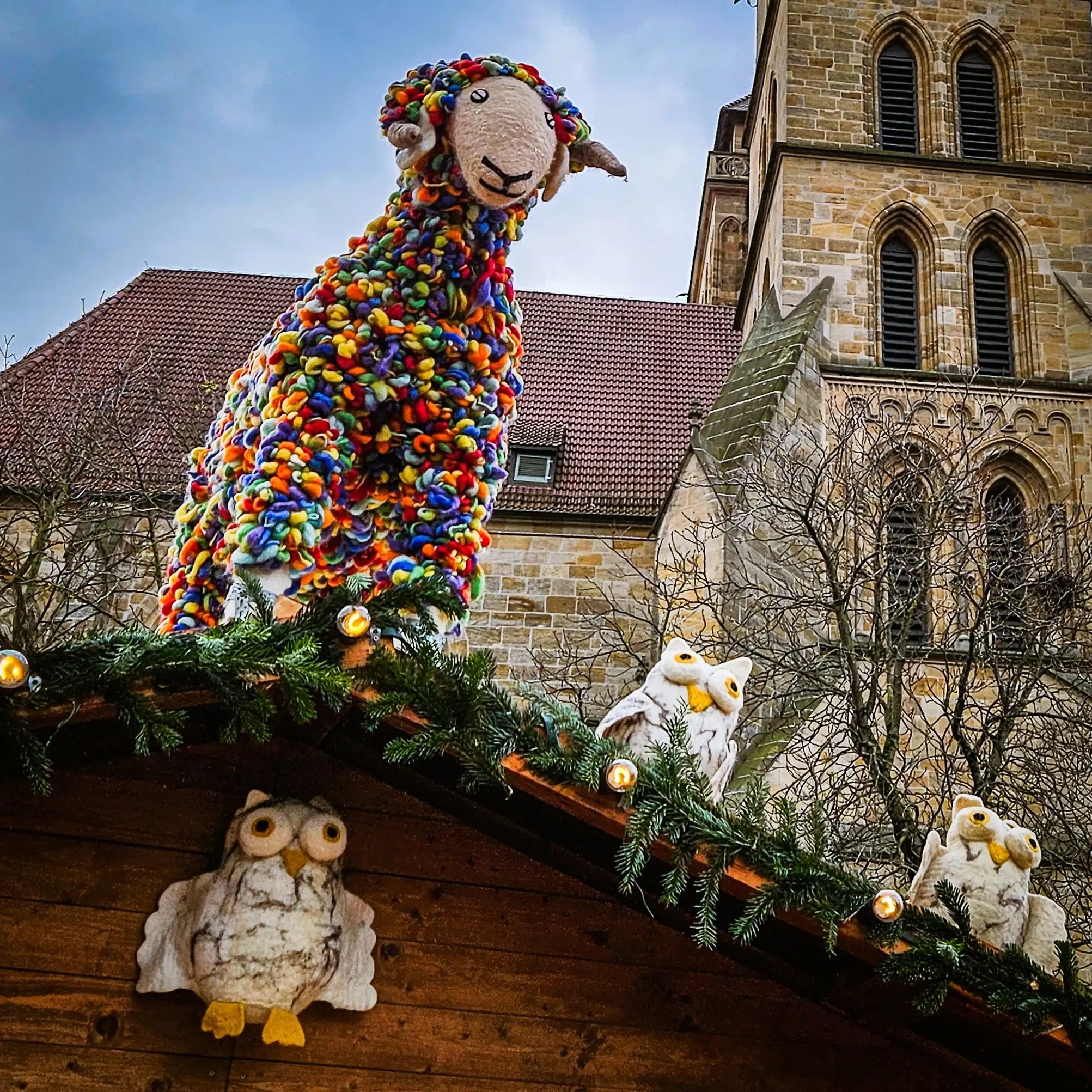 A large rainbow-colored sheep decoration and felt owls on the roof of a Christmas market stall in front of St. Dionys Church.