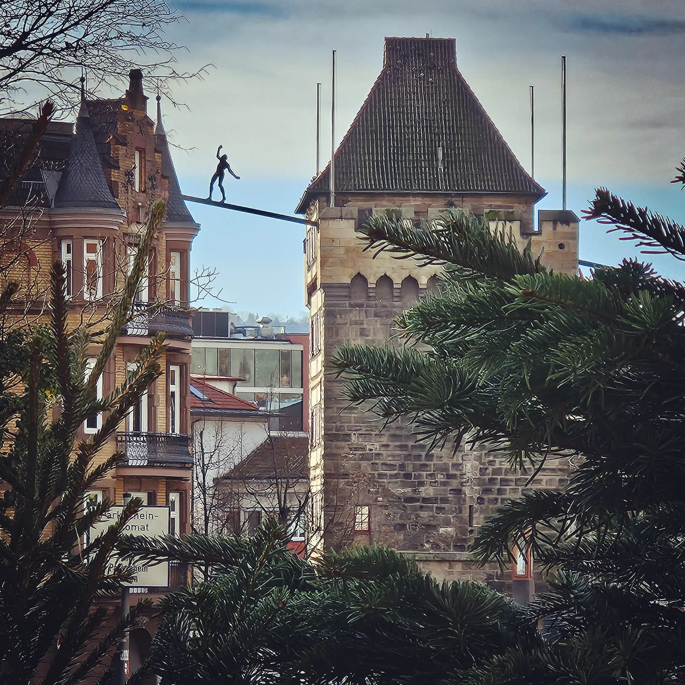 A metal tightrope walker sculpture balanced between two historic towers in Esslingen, framed by evergreen branches.