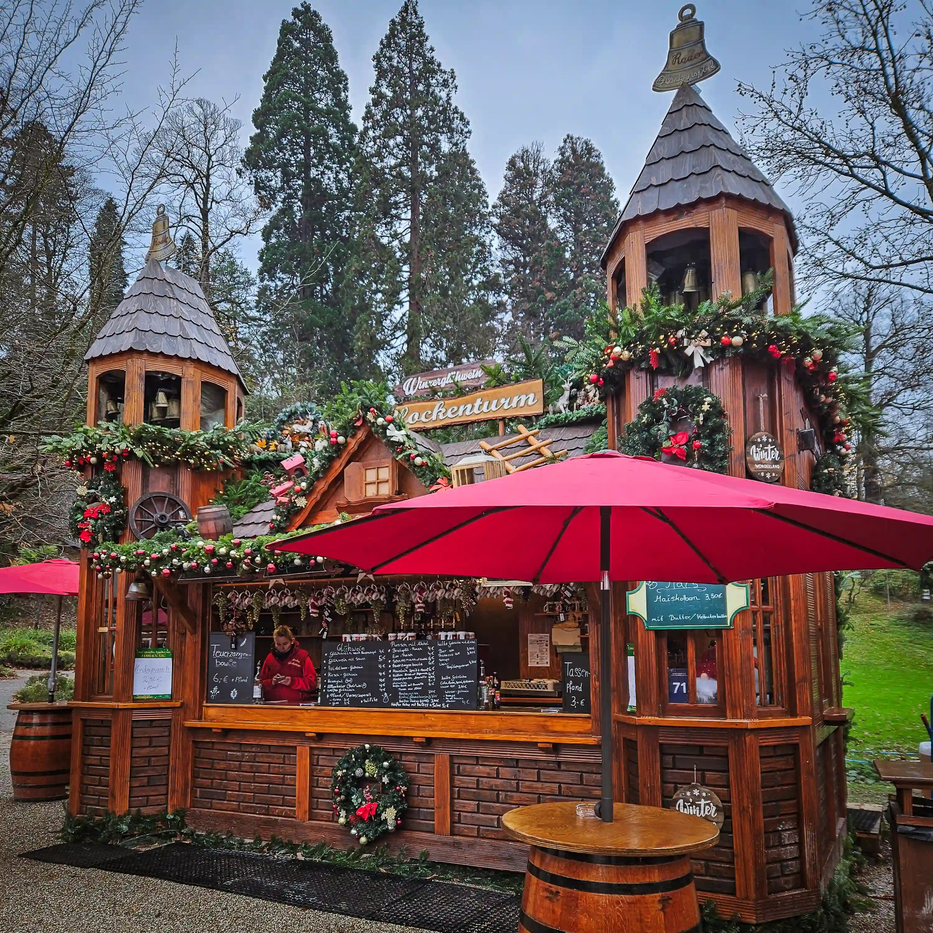 Wooden stall with two small towers, festive garlands, and a red umbrella in front.