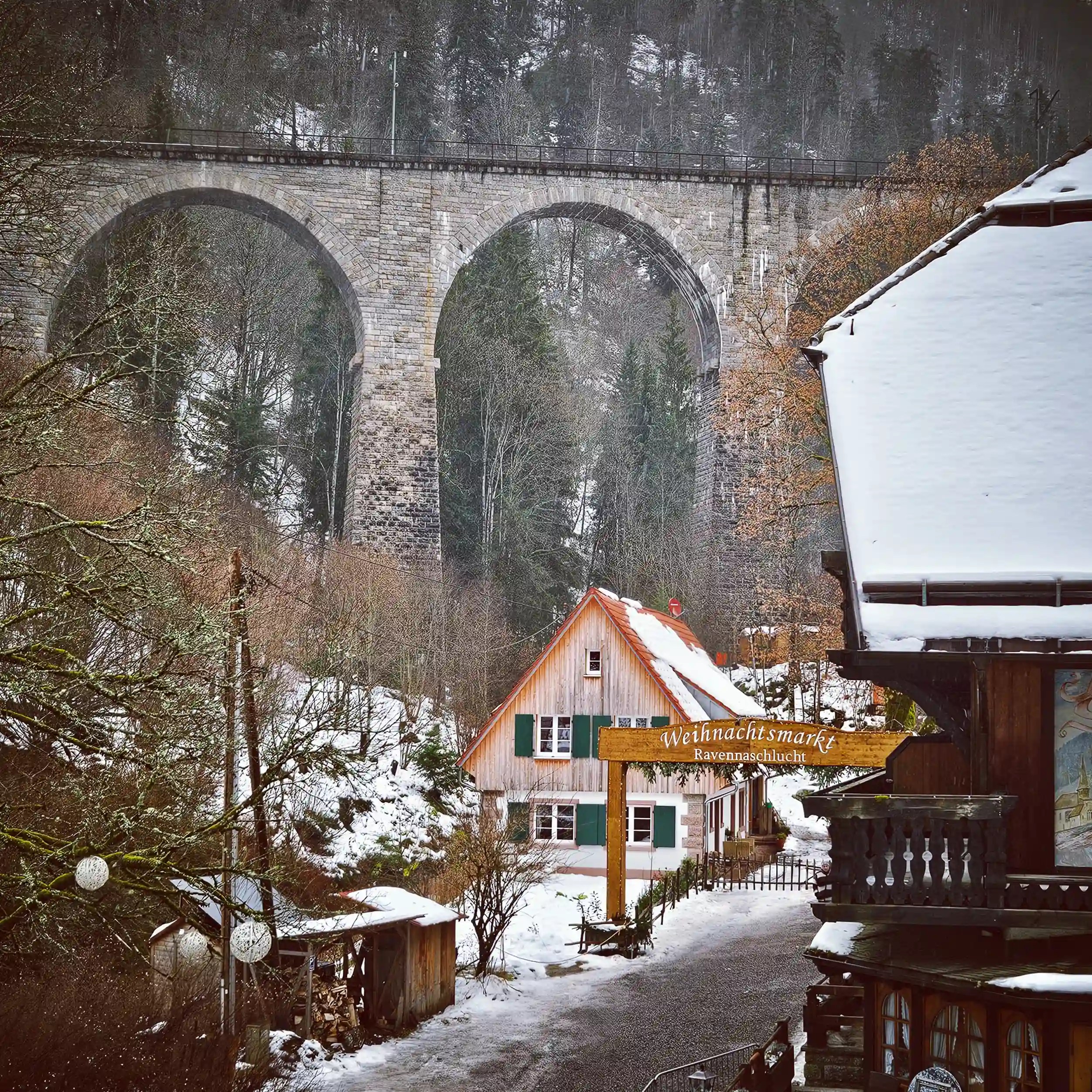Snowy view of the stone Ravenna Viaduct towering above a wooden chalet and sign reading “Weihnachtsmarkt Ravennaschlucht.”
