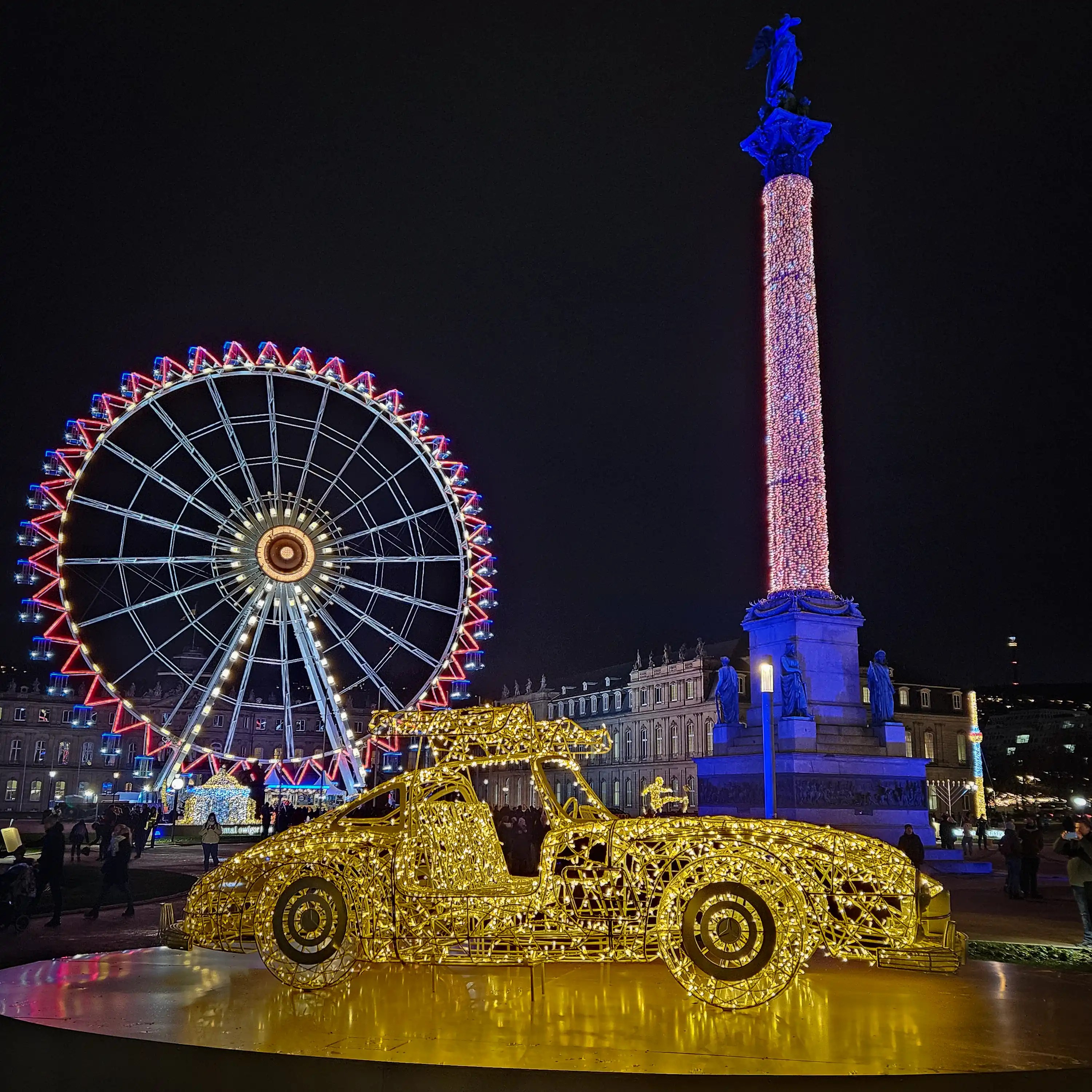 Large illuminated Ferris wheel and golden car sculpture in Stuttgart’s Schlossplatz at night.