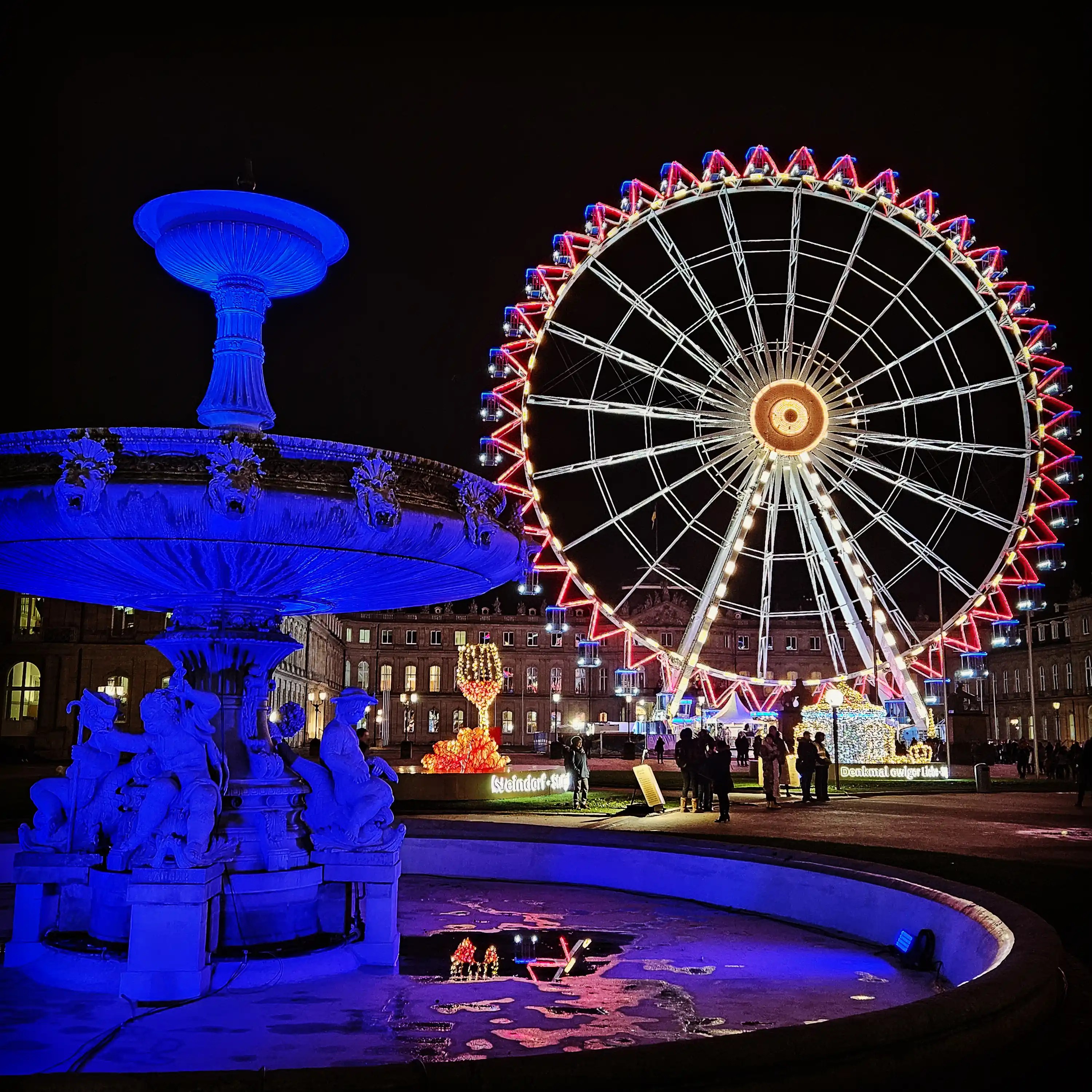 Fountain and Ferris wheel illuminated in blue and red lights at Stuttgart’s Schlossplatz.