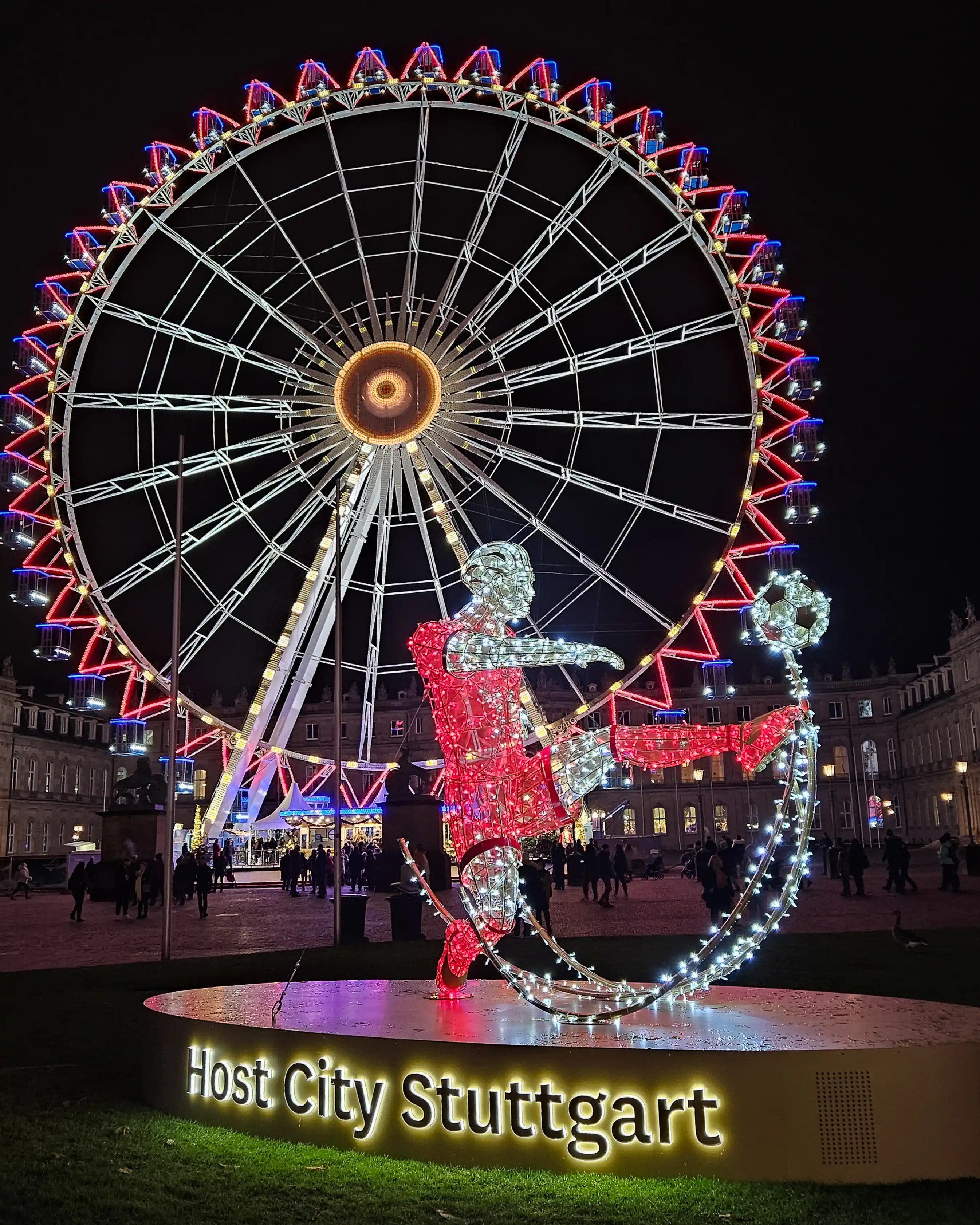 Illuminated figure of a soccer player kicking a ball in front of Stuttgart’s Ferris wheel, labeled “Host City Stuttgart.”