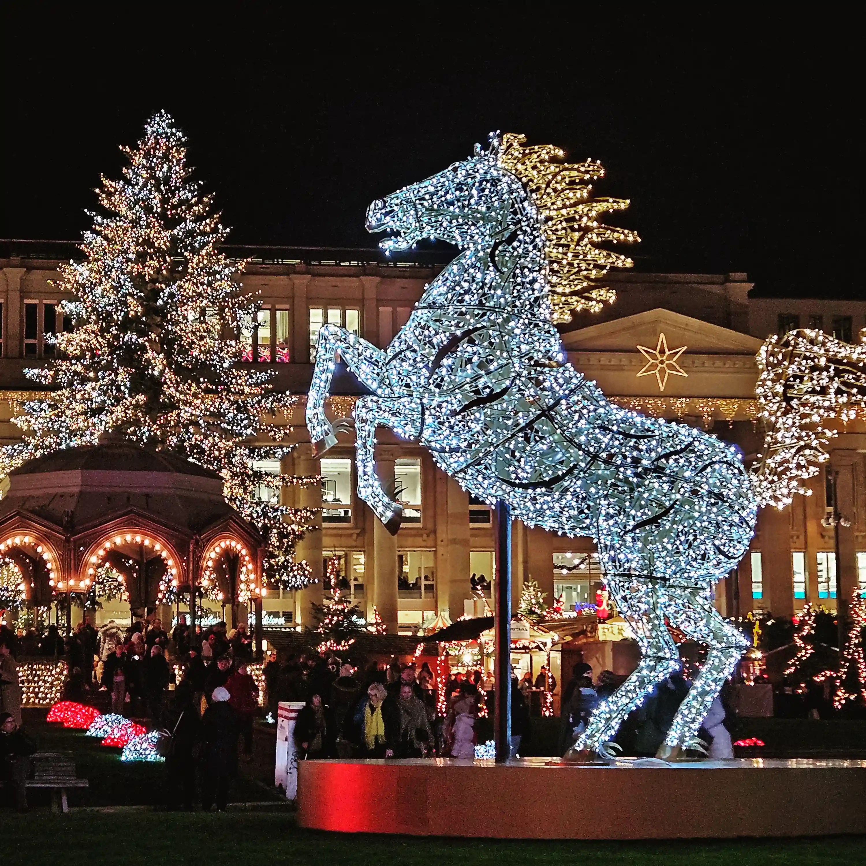 Large illuminated horse sculpture and Christmas tree surrounded by market stalls and lights in Stuttgart’s Schlossplatz.