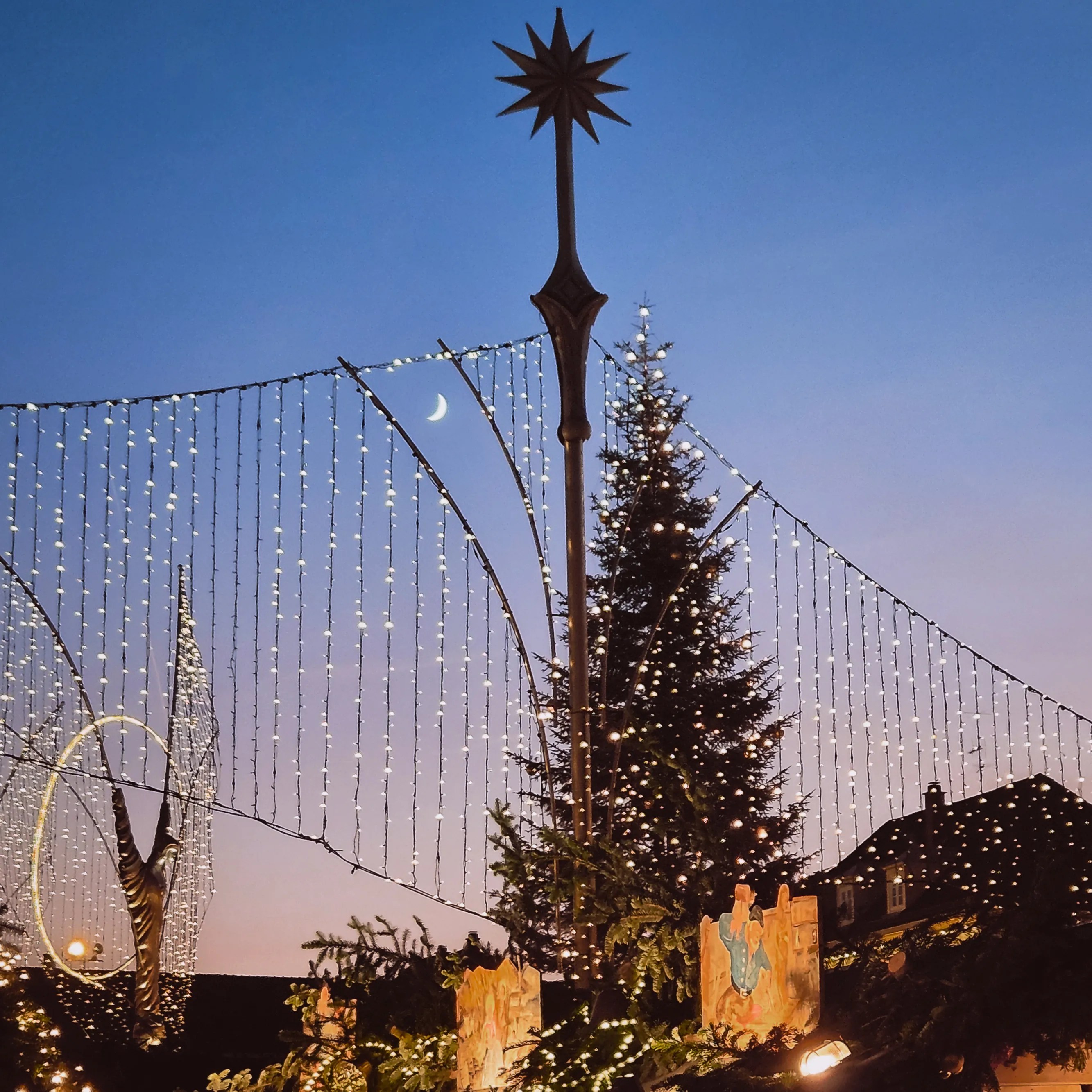 Crescent moon rising above twinkling lights, Christmas trees, and decorations at the Ludwigsburg Baroque Christmas Market at dusk.