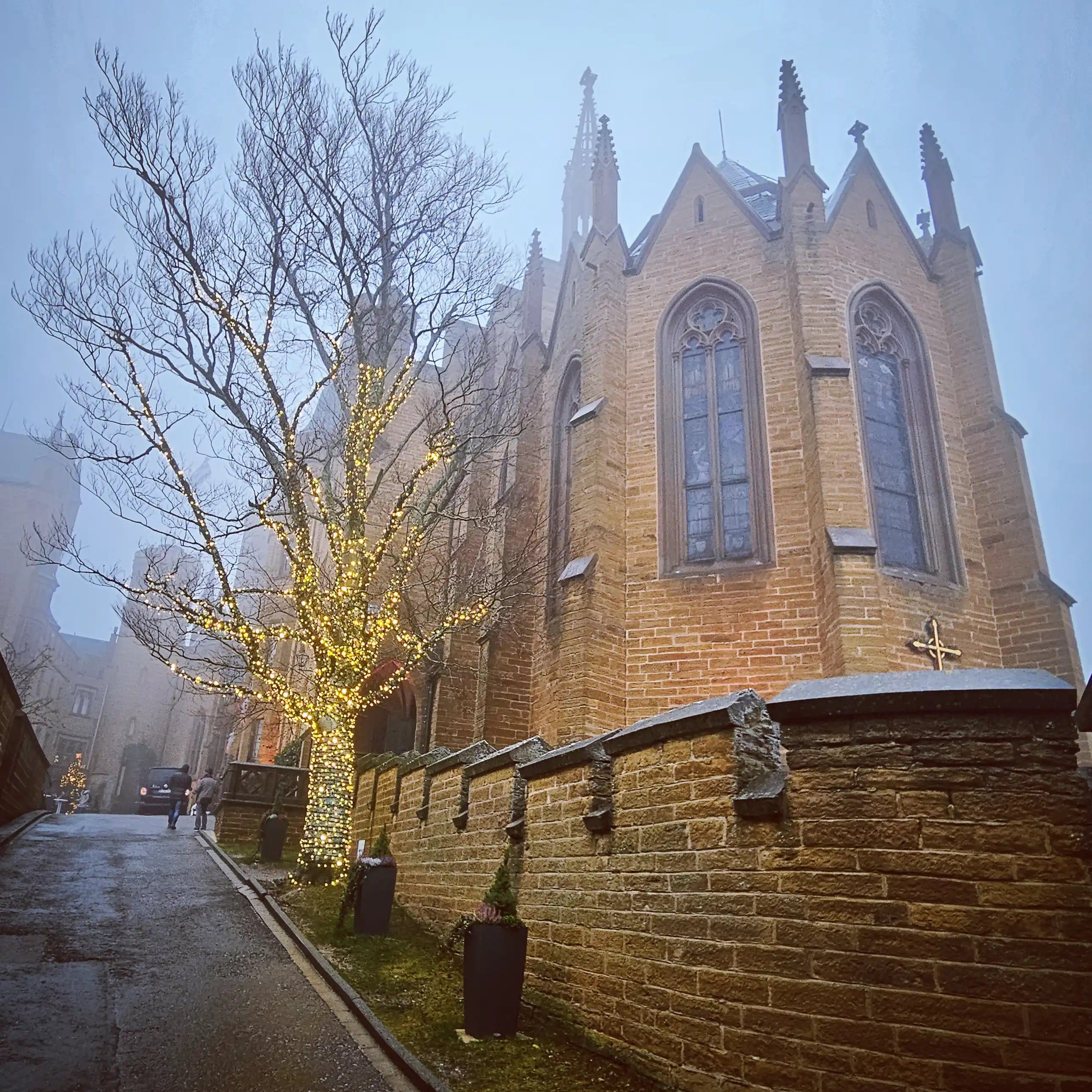 Tree wrapped in fairy lights beside the Gothic chapel at Burg Hohenzollern on a foggy evening.