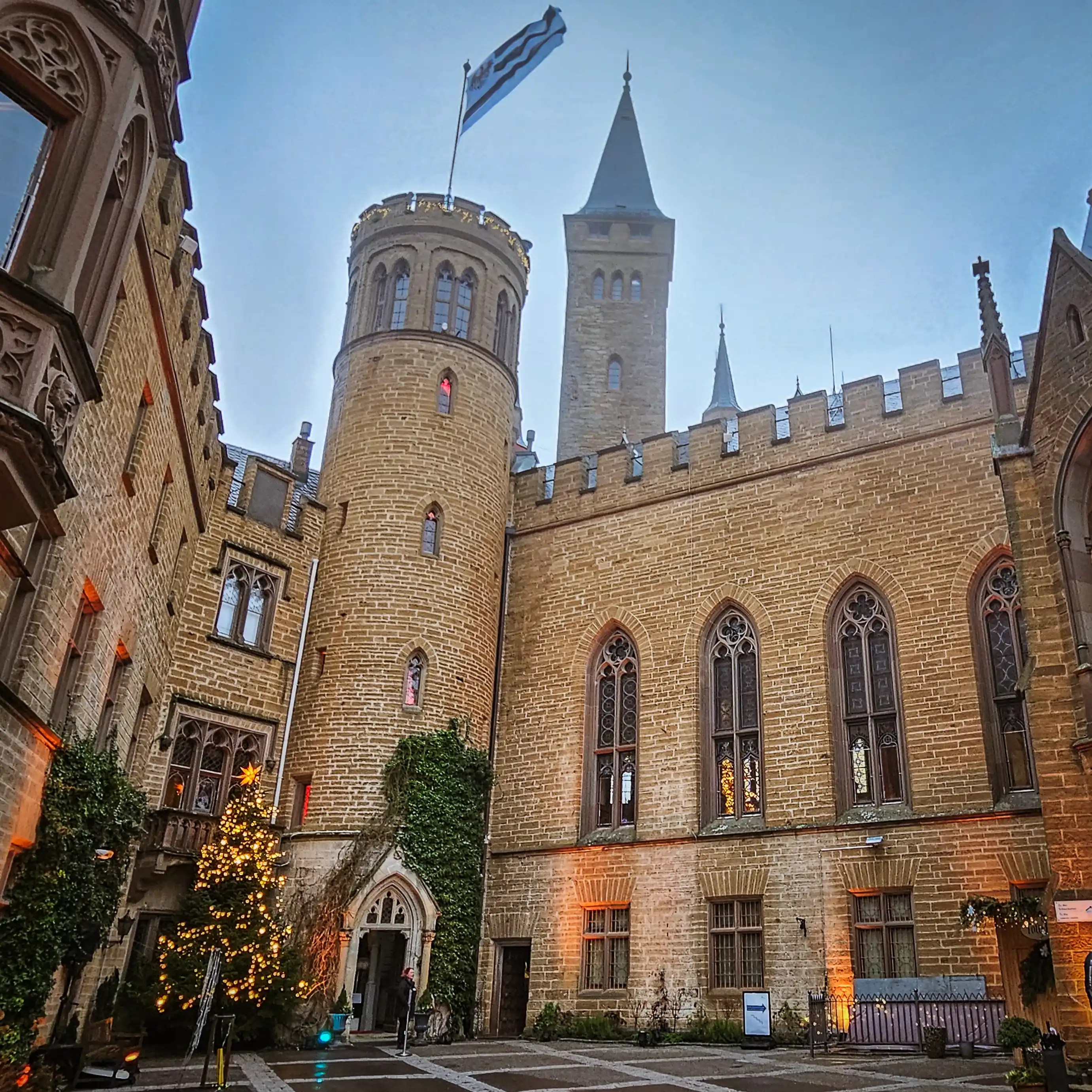 Courtyard of Burg Hohenzollern with festive lights, Christmas tree, and flying flag on a misty day.