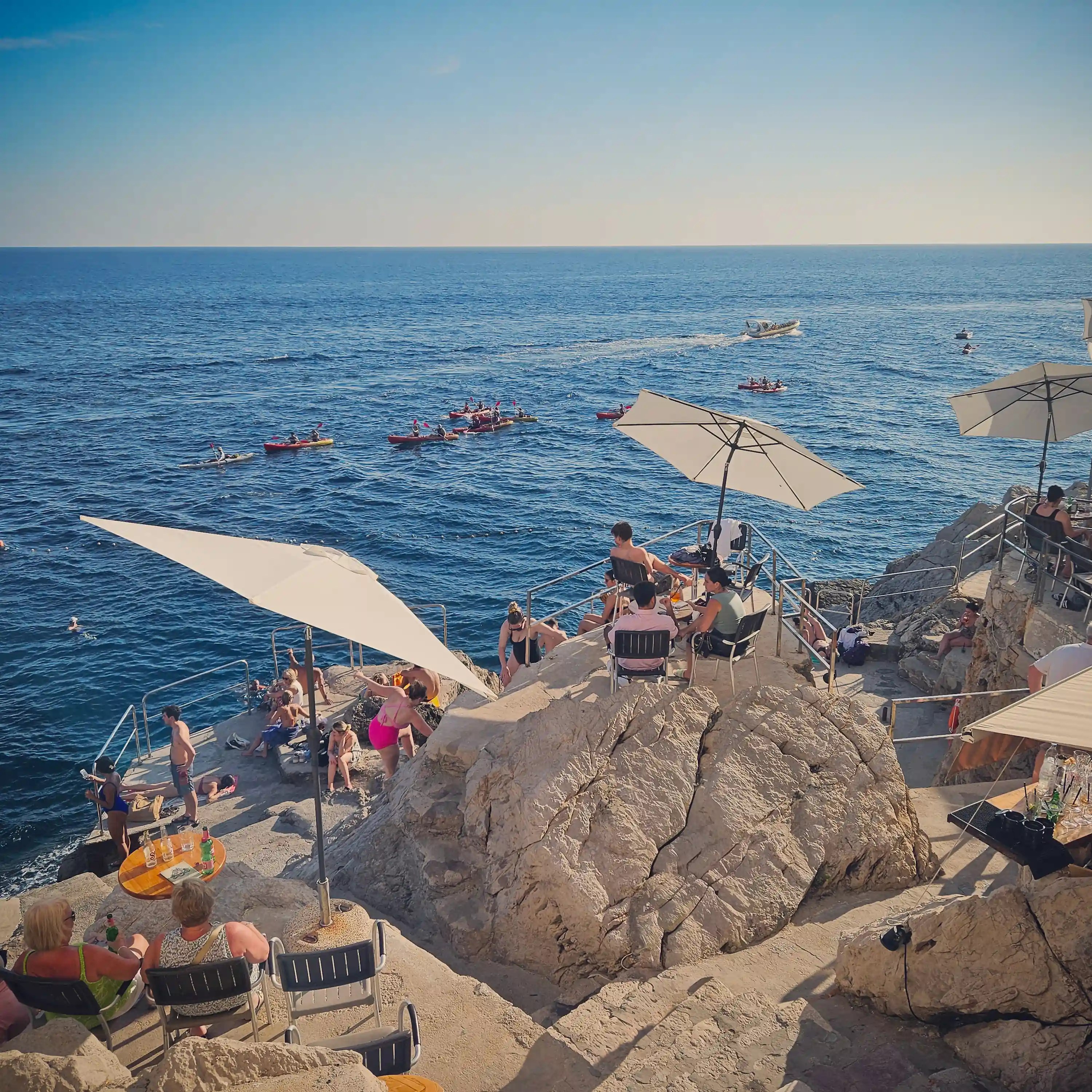 People relaxing under umbrellas on rocky cliffs above the Adriatic Sea in Dubrovnik.