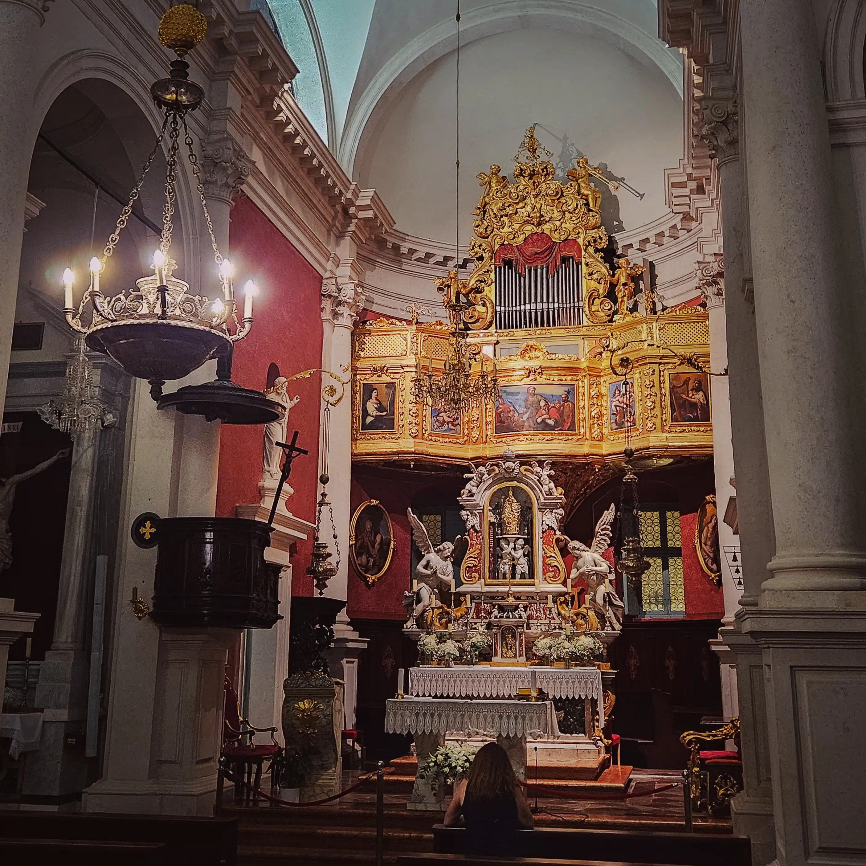 Interior of St. Blaise Church in Dubrovnik, Croatia, showing the ornate Baroque altar with golden details, angel statues, and a visitor seated in quiet reflection.