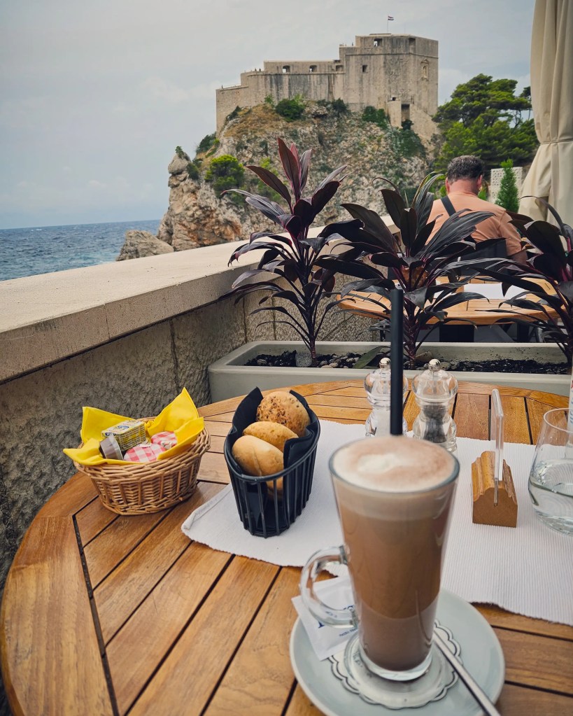 Latte and basket of rolls on a seaside café table overlooking Dubrovnik’s fortress.