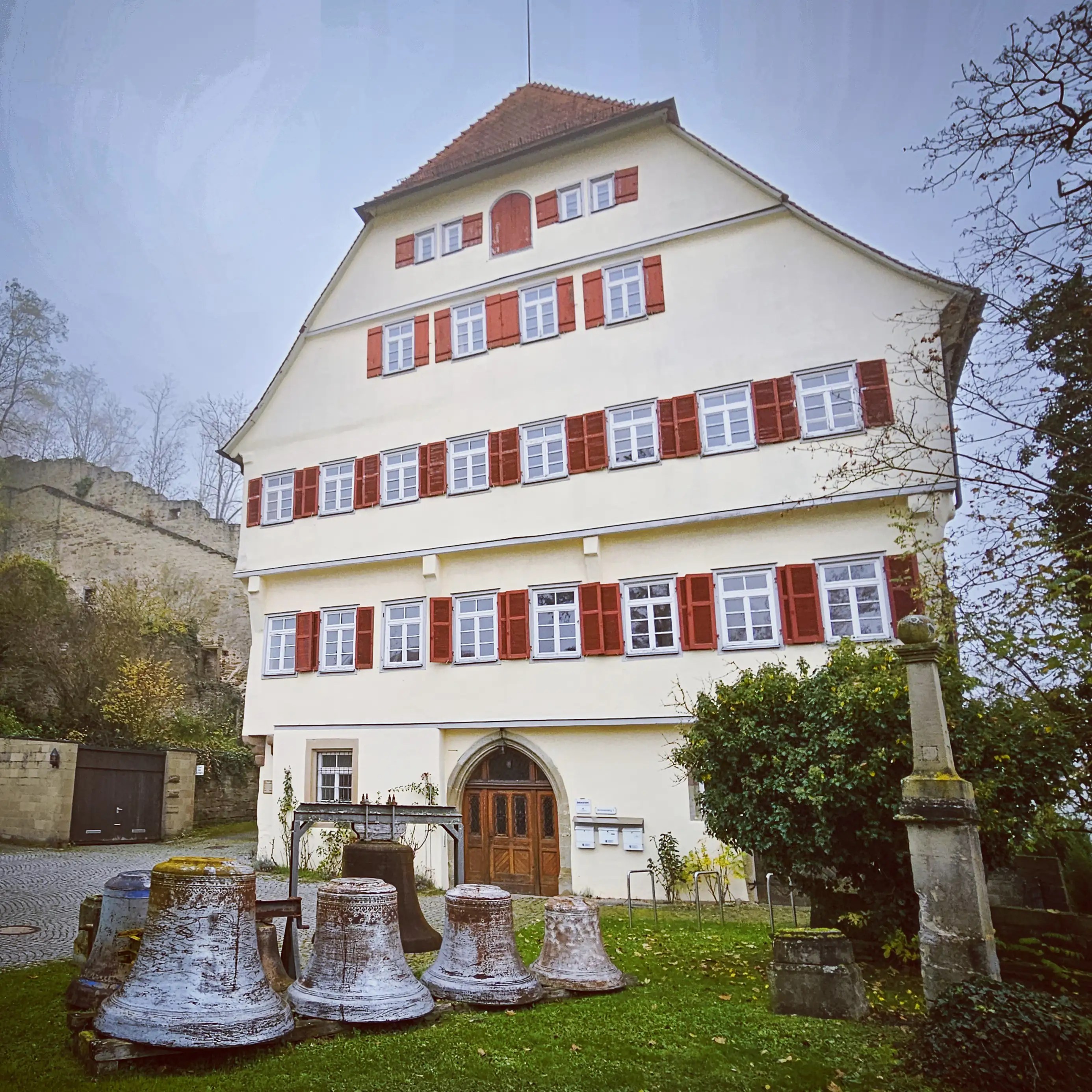 Large white building with red shutters beside old church bells in Herrenberg, Germany.