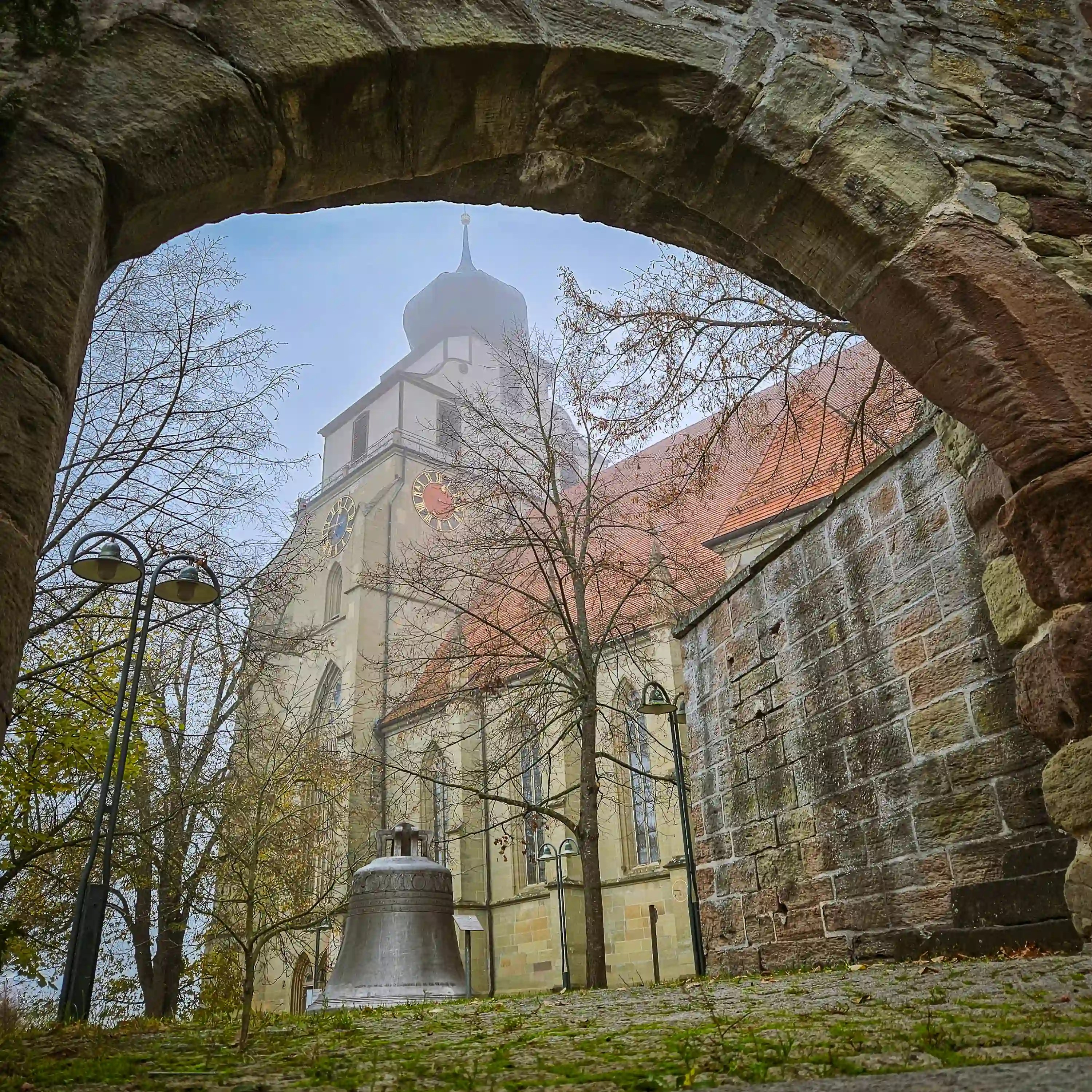 View of Stiftskirche Herrenberg through a stone archway with a large bell in front.