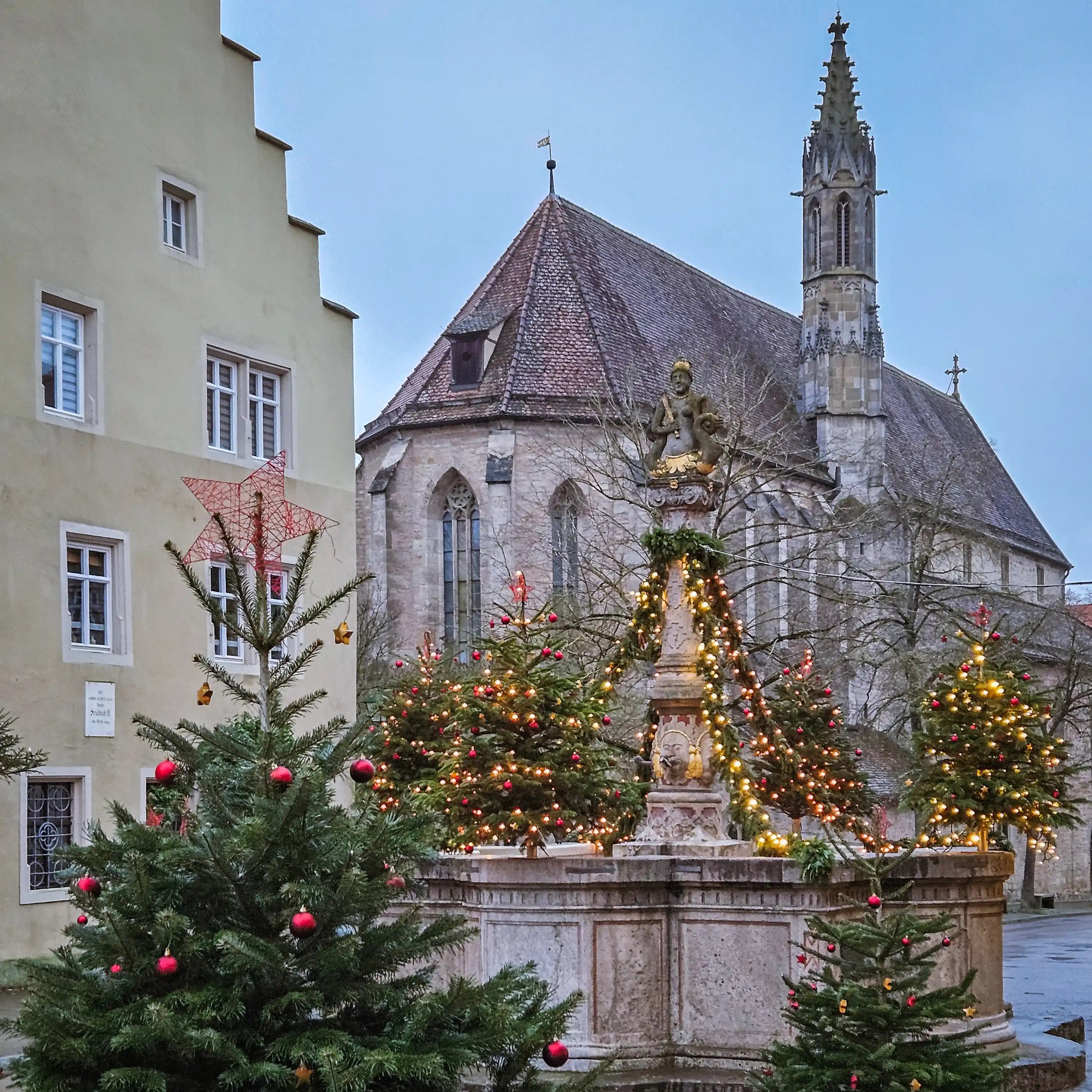 Christmas trees and lights surrounding the Herrnbrunnen Fountain with the Franciscan Church in the background in Rothenburg ob der Tauber.