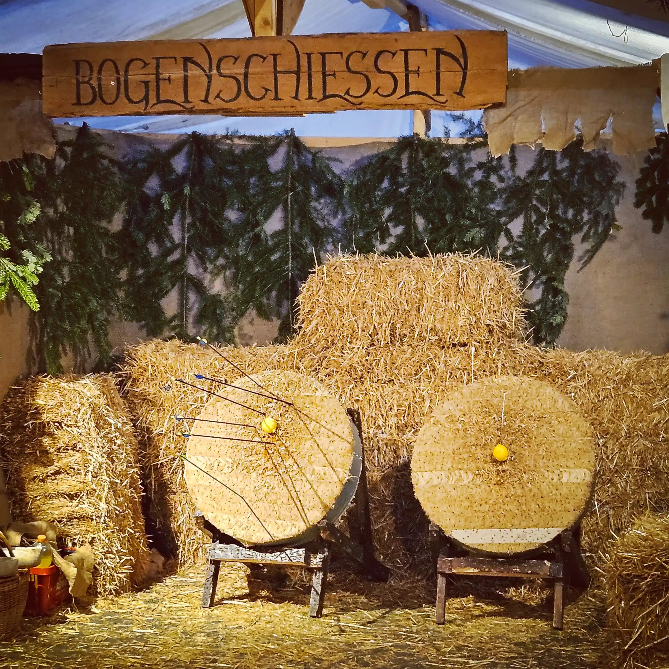 A medieval-style archery range with straw targets and a wooden sign reading “Bogenschiessen.”