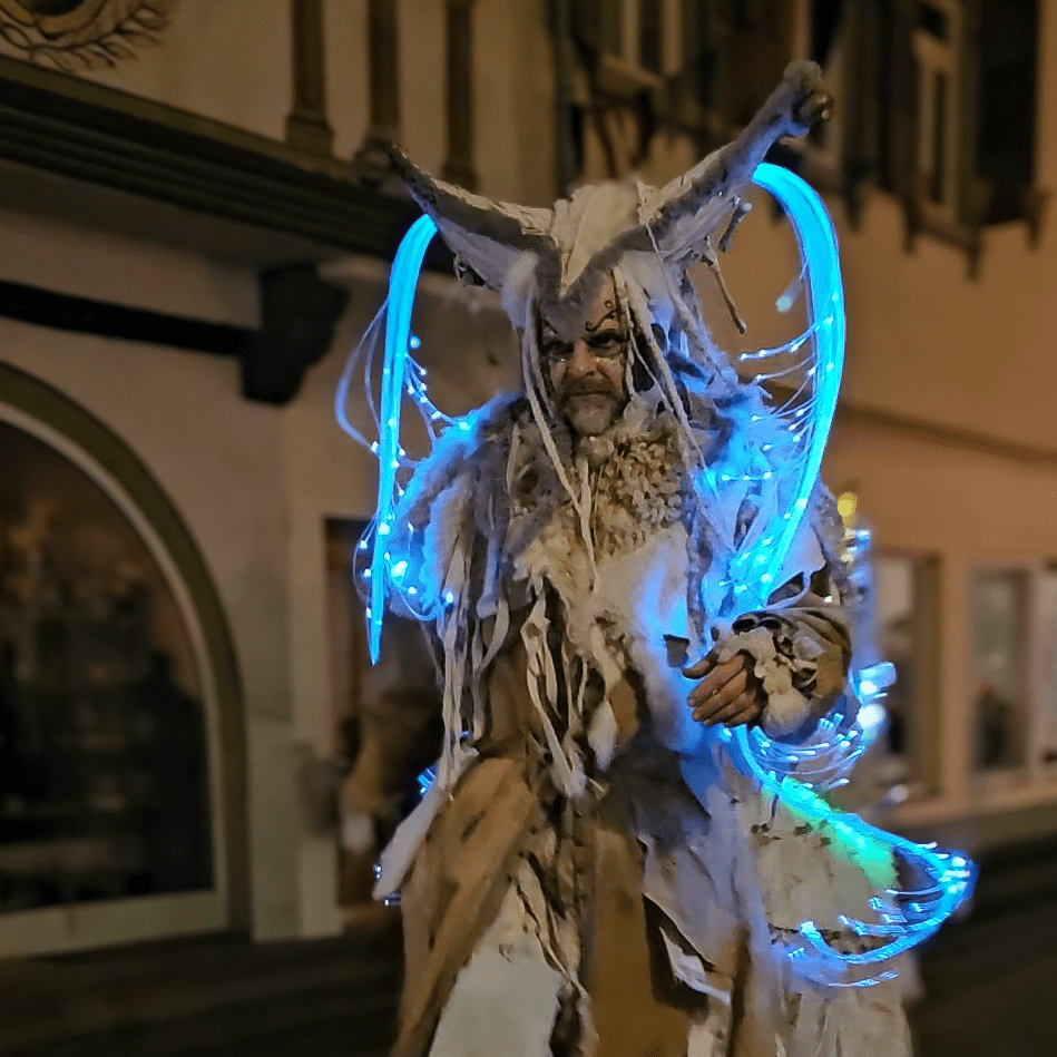 A costumed performer in fur and glowing blue lights walks through the Esslingen Medieval Christmas Market at night.
