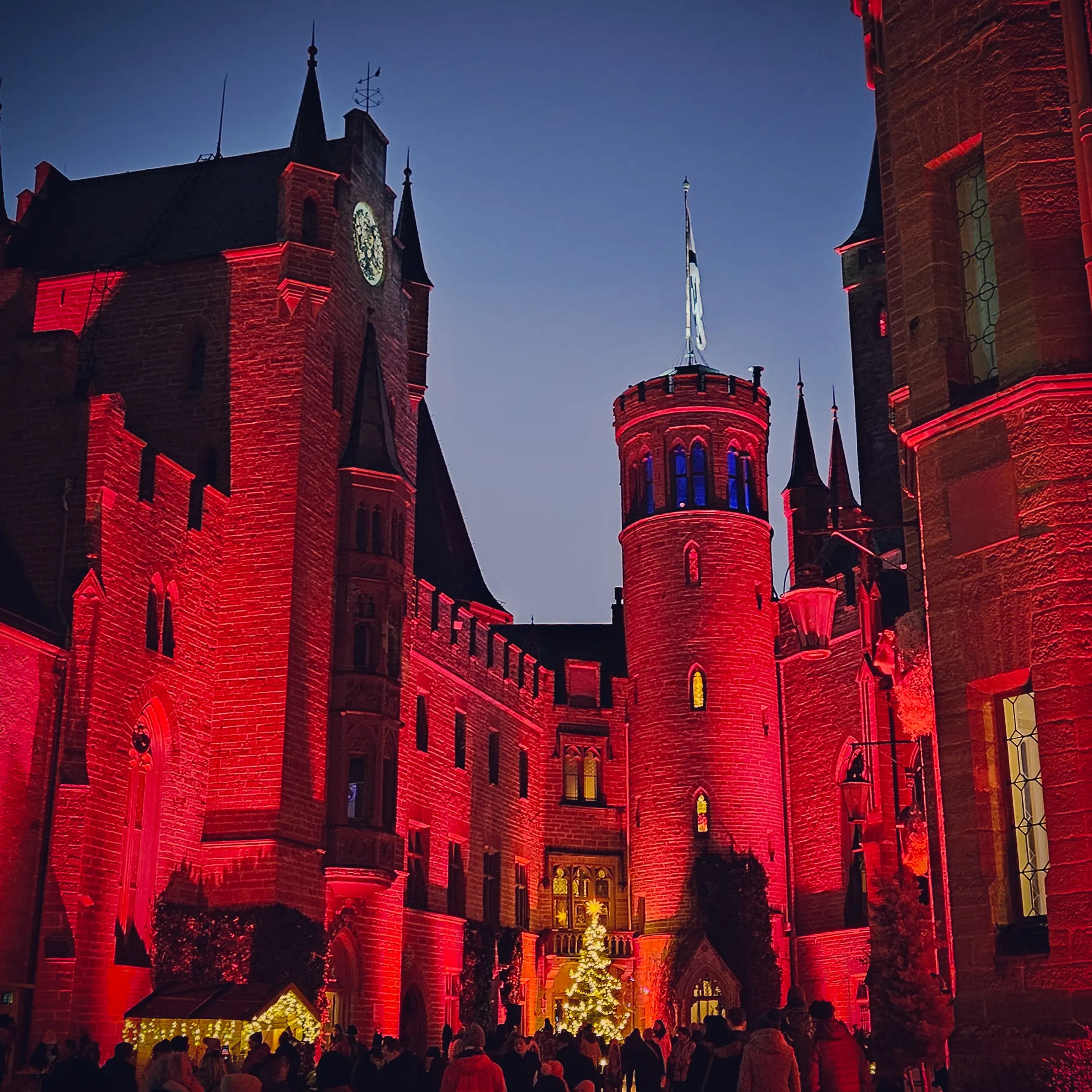 The courtyard of Burg Hohenzollern illuminated in red light with visitors gathered around a Christmas tree at dusk.