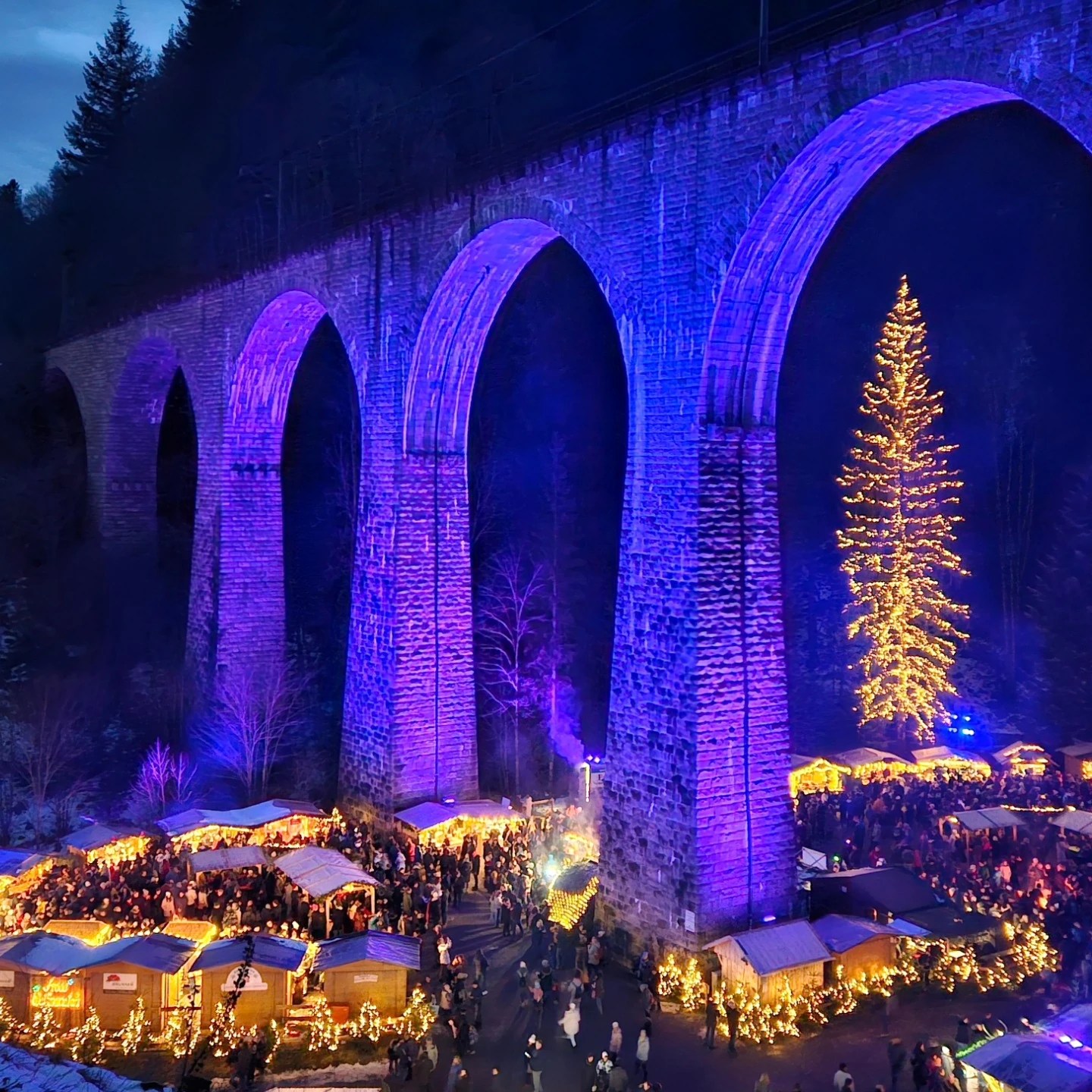 Aerial view of the Ravenna Gorge Christmas Market illuminated beneath the viaduct, with glowing stalls and a tall Christmas tree.