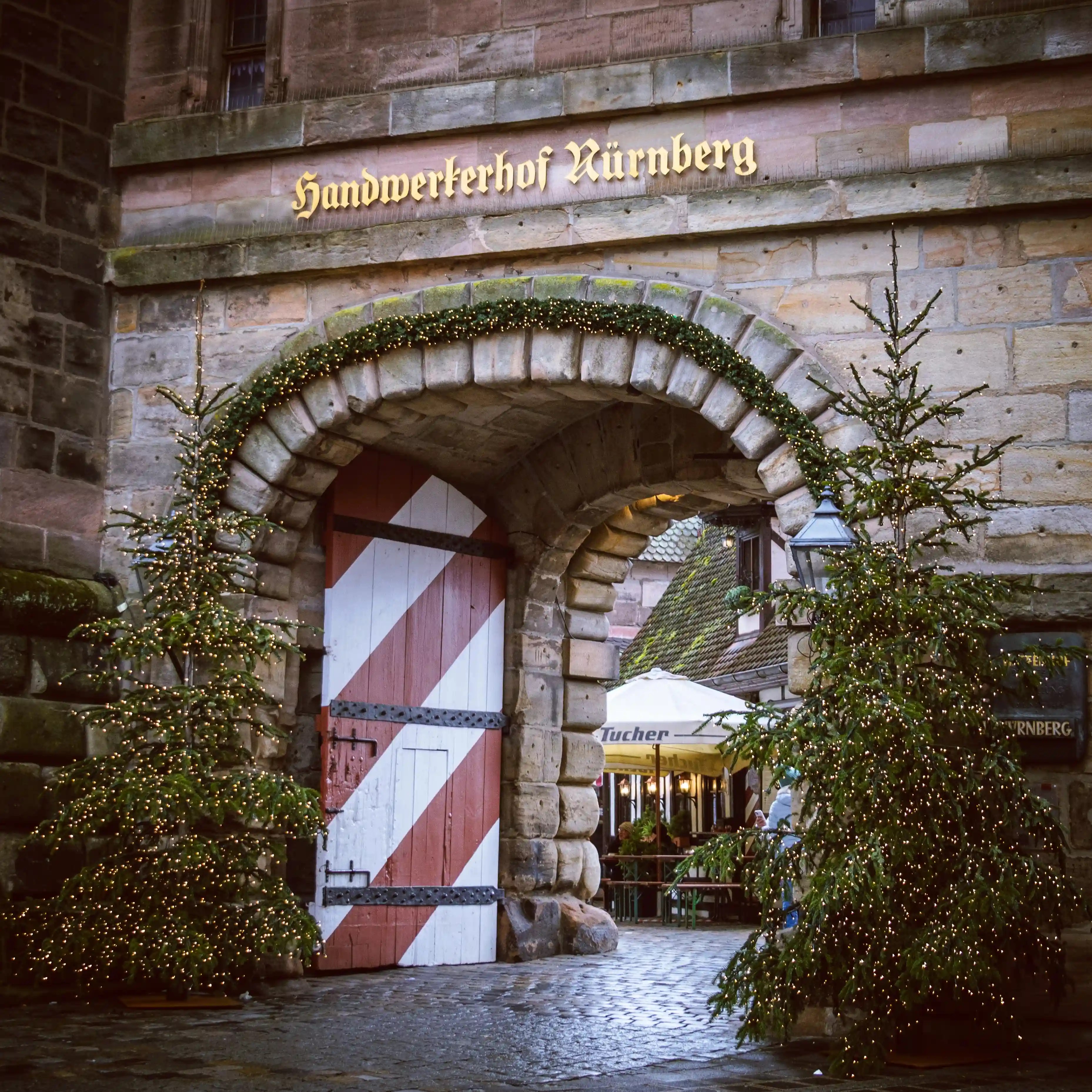 Arched stone gate of the Handwerkerhof Nürnberg decorated with twinkling lights and Christmas trees.