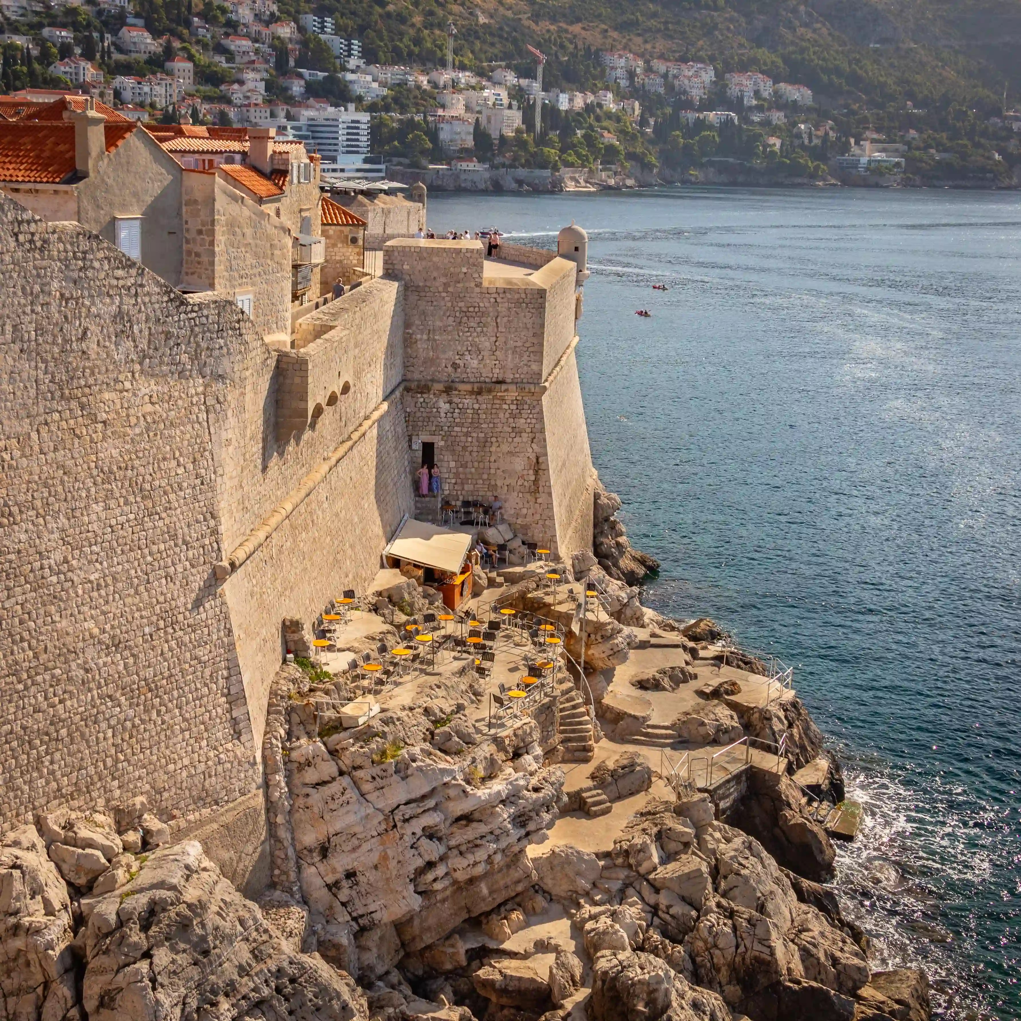 View of Bard Dubrovnik, a cliffside bar built just outside Dubrovnik’s city walls, with yellow tables and chairs set among rocky ledges overlooking the Adriatic Sea.