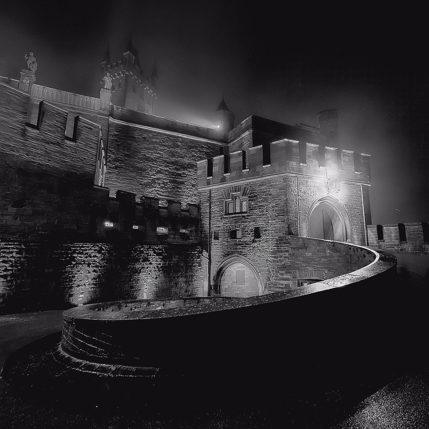 Black and white photograph of Burg Hohenzollern’s illuminated ramparts and arched entryway shrouded in mist.