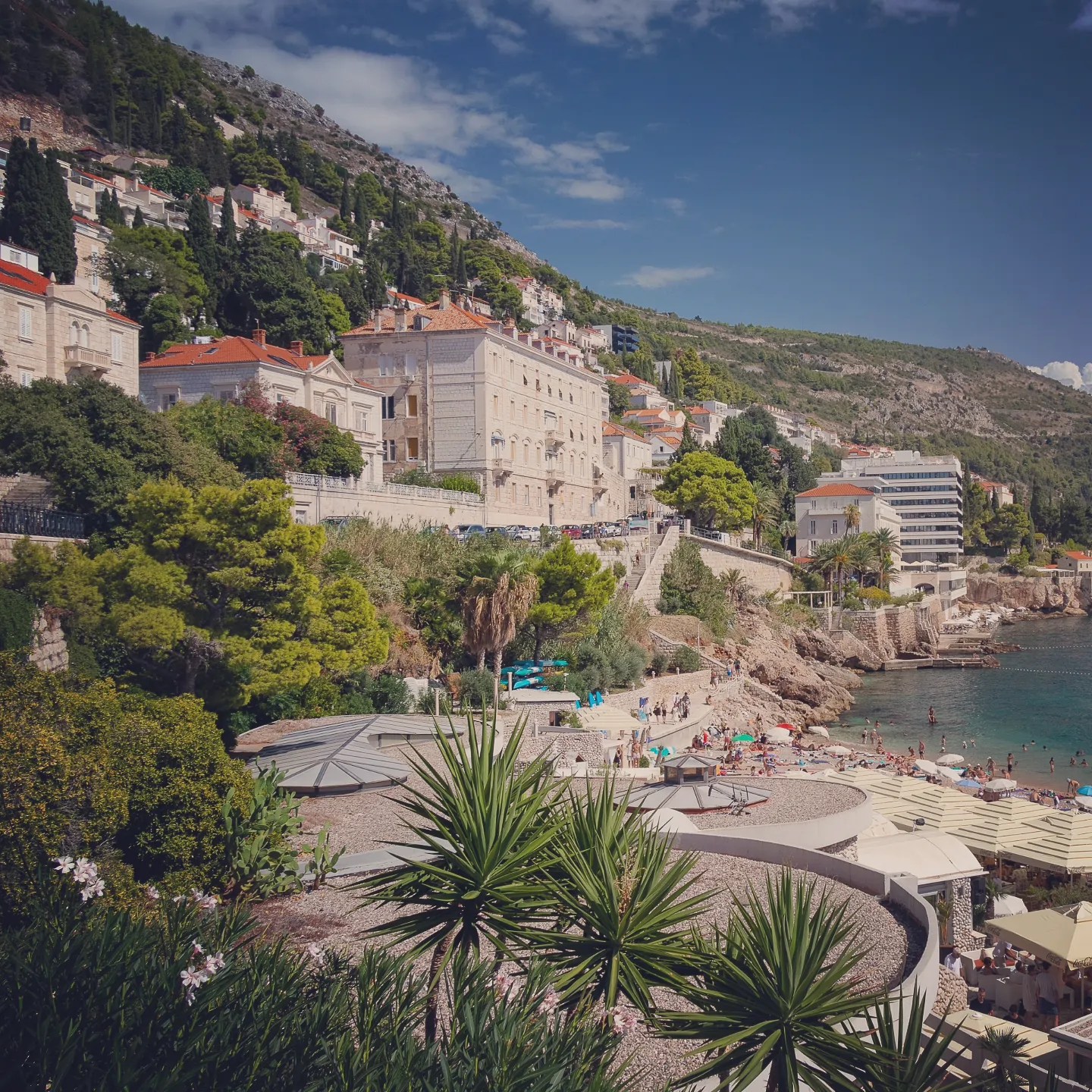 Banje Beach in Dubrovnik with sunbathers, umbrellas, and hillside villas overlooking the Adriatic Sea.
