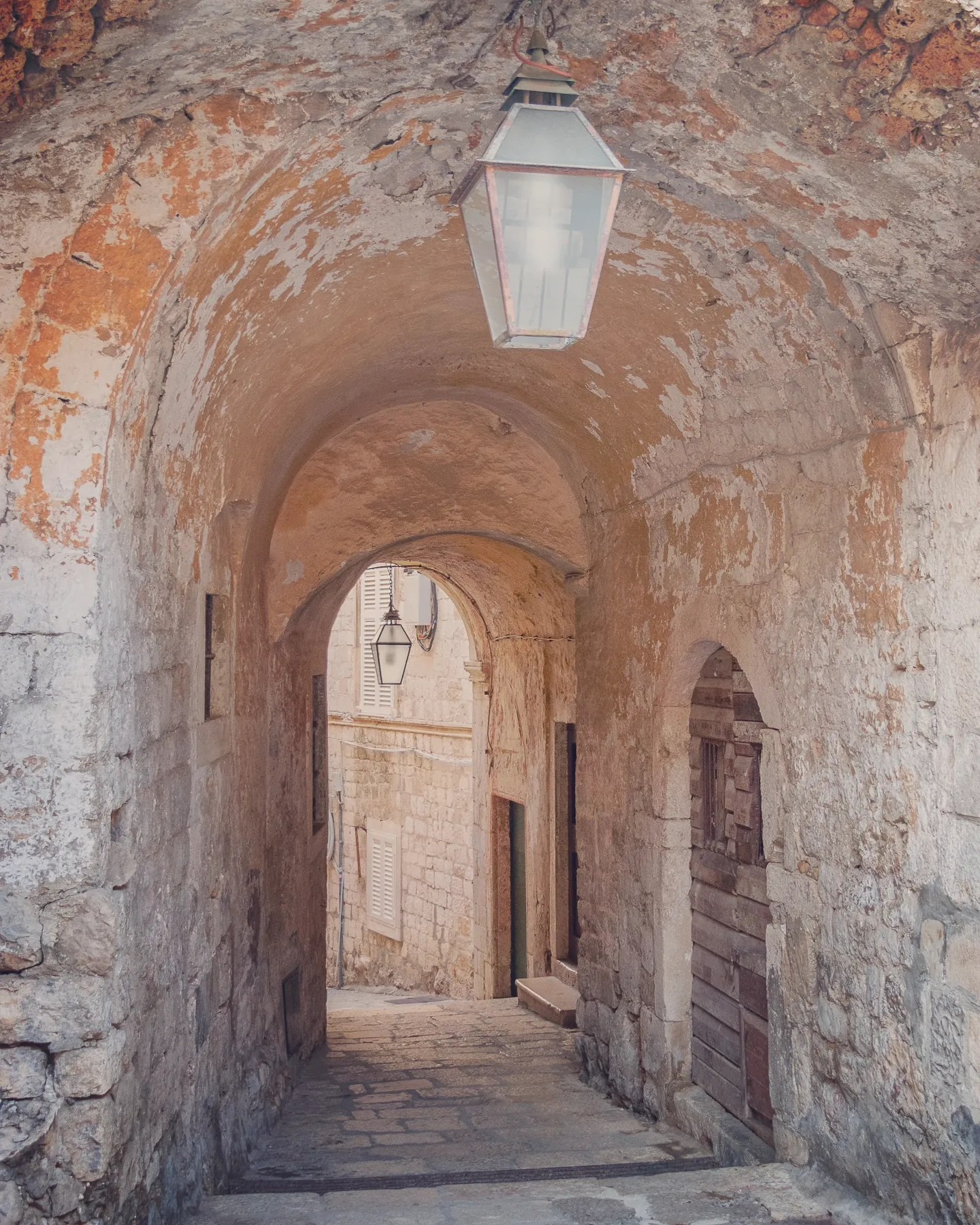 Narrow stone passage with weathered arches and hanging lanterns in Dubrovnik’s Old Town.