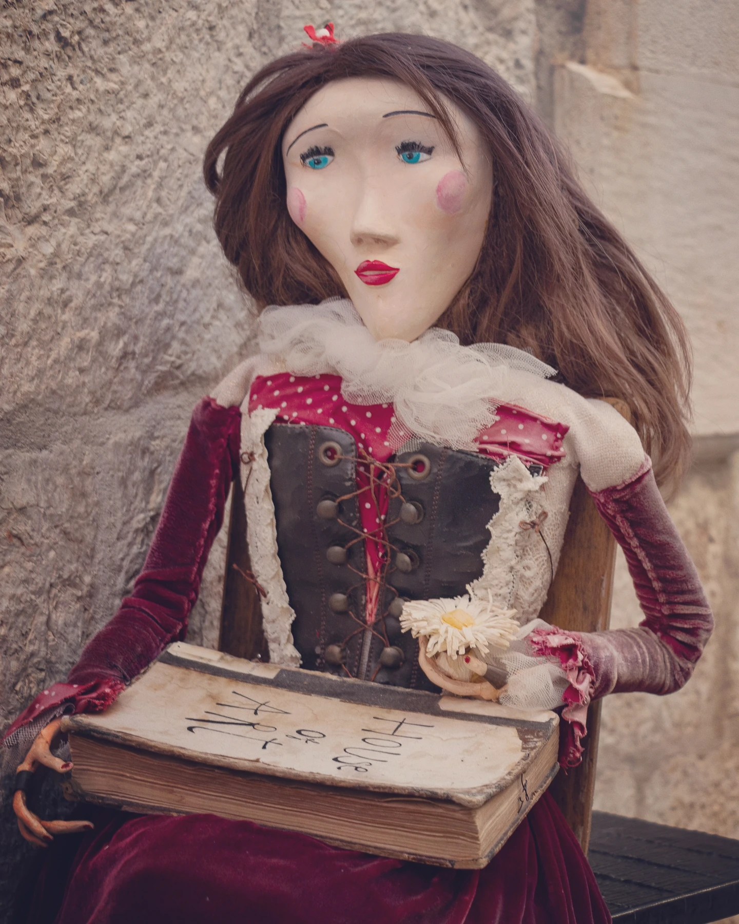 A handmade doll in a red velvet dress holding a book and flower, displayed outside a shop in Dubrovnik.