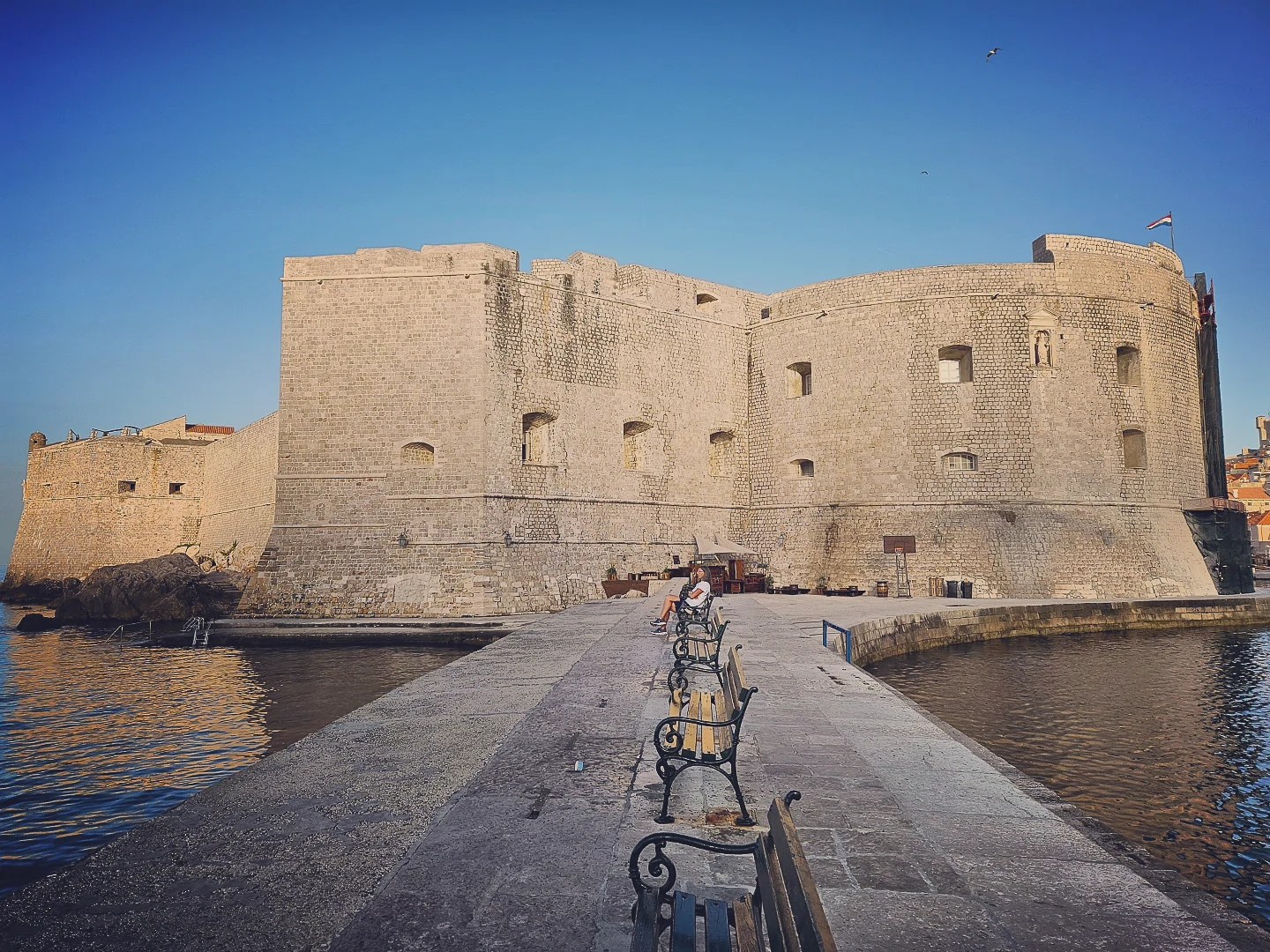 Stone walls of Fort St. John in Dubrovnik with benches along the harbor walkway at sunrise.
