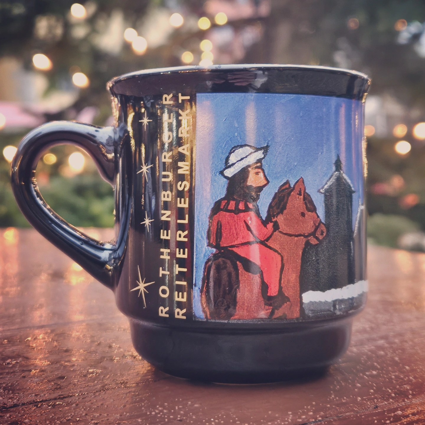 Close-up of a Rothenburg Reiterlesmarkt mug featuring a rider on horseback, with blurred Christmas lights in the background.