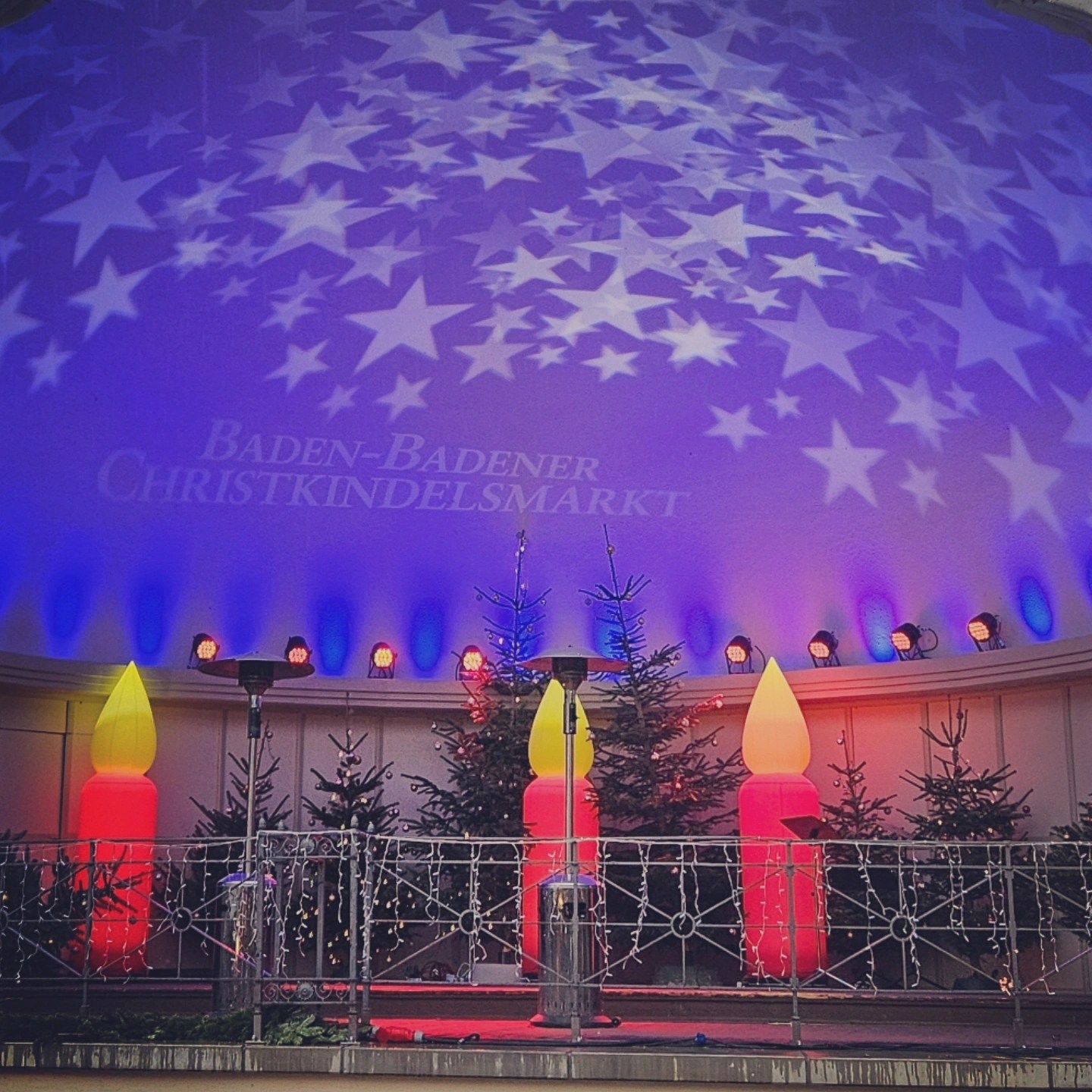 Stage at the Baden-Badener Christkindelsmarkt illuminated with stars and colorful lights, decorated with Christmas trees and candle displays.